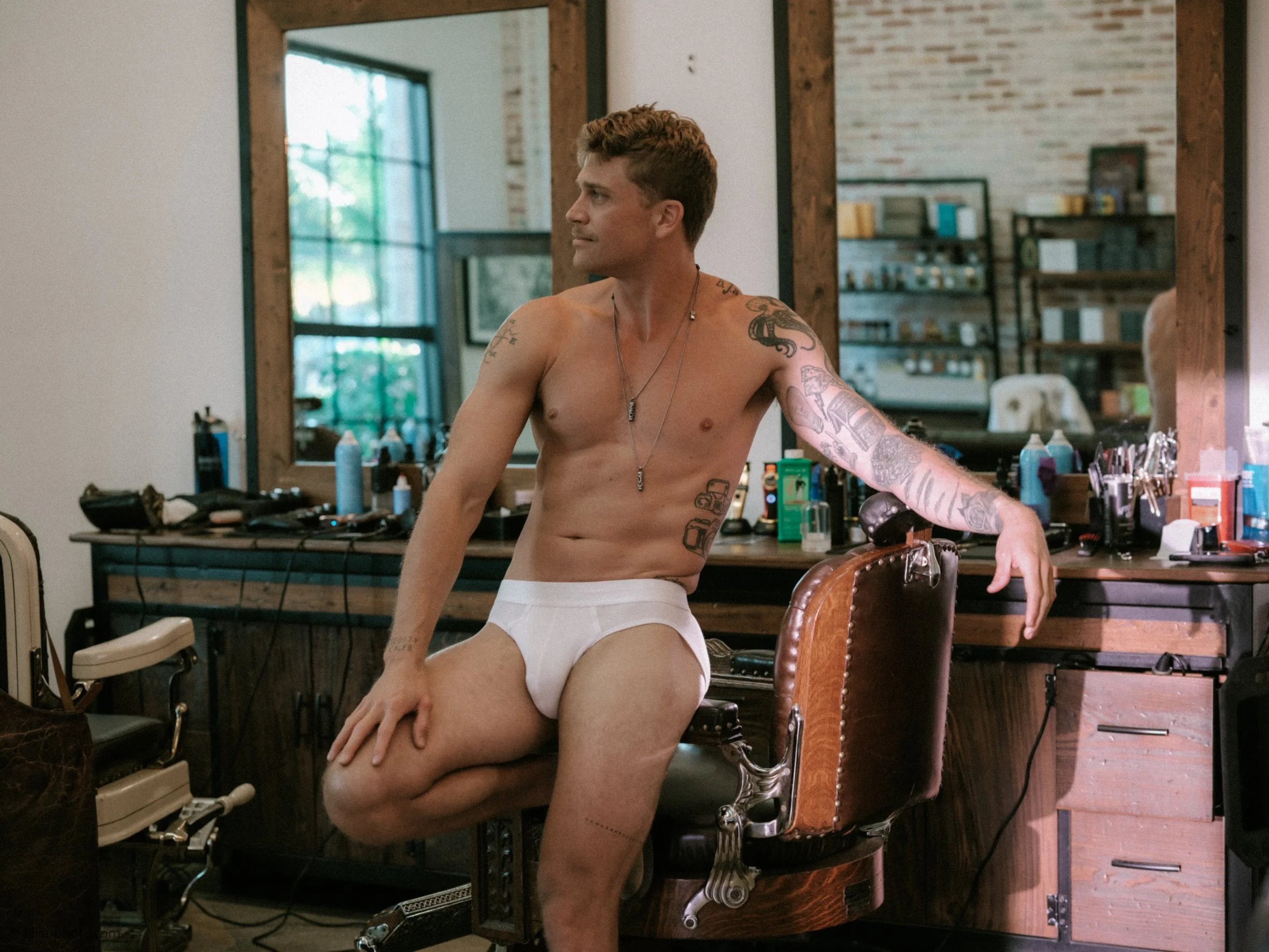 A shirtless man with tattoos sitting on a barber chair in a barbershop, with grooming products and tools on the counter behind him, and a large mirror reflecting the shop's interior.