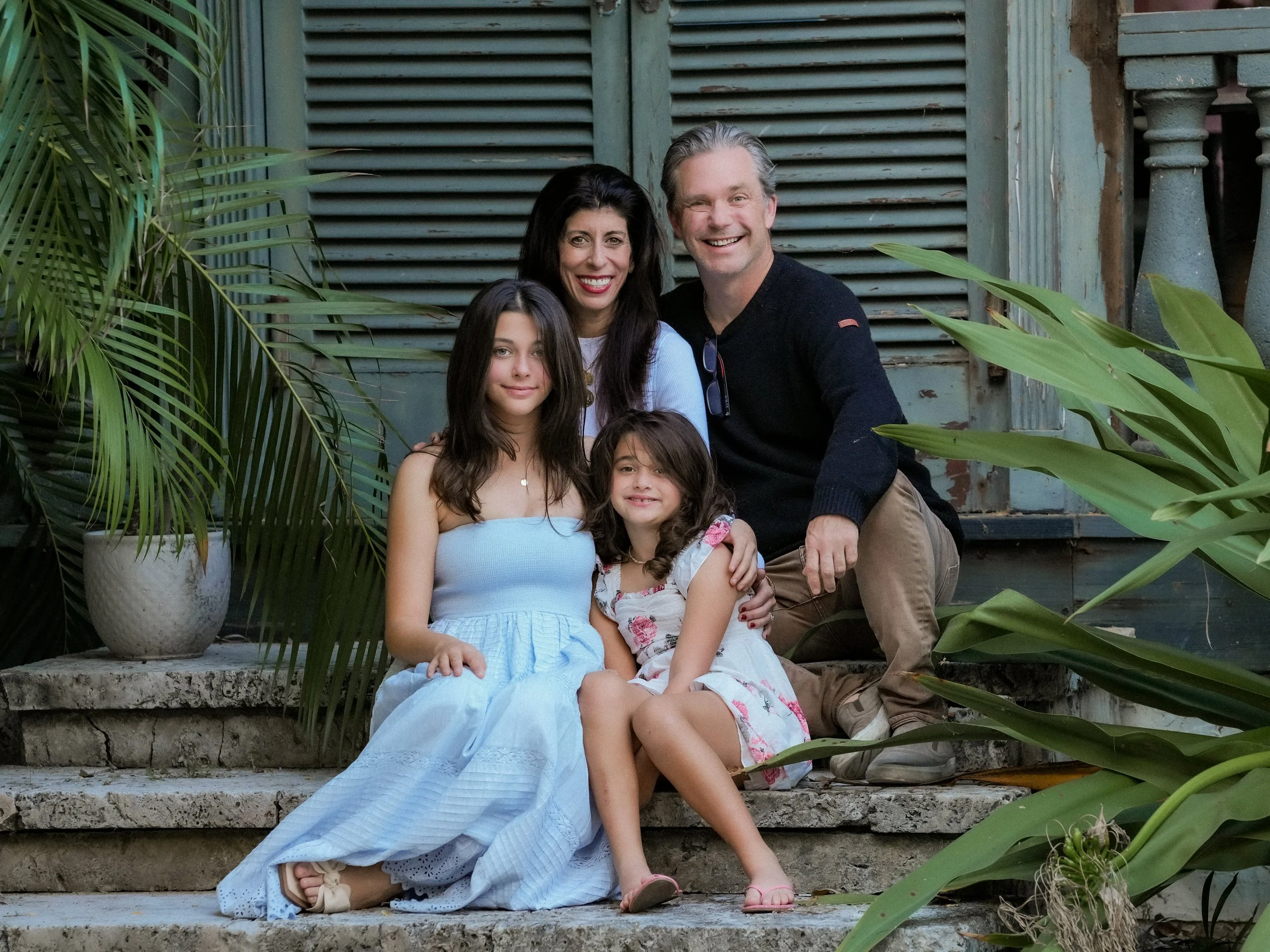 A family of four posed outdoors on stone steps, surrounded by greenery, with rustic shutters in the background.