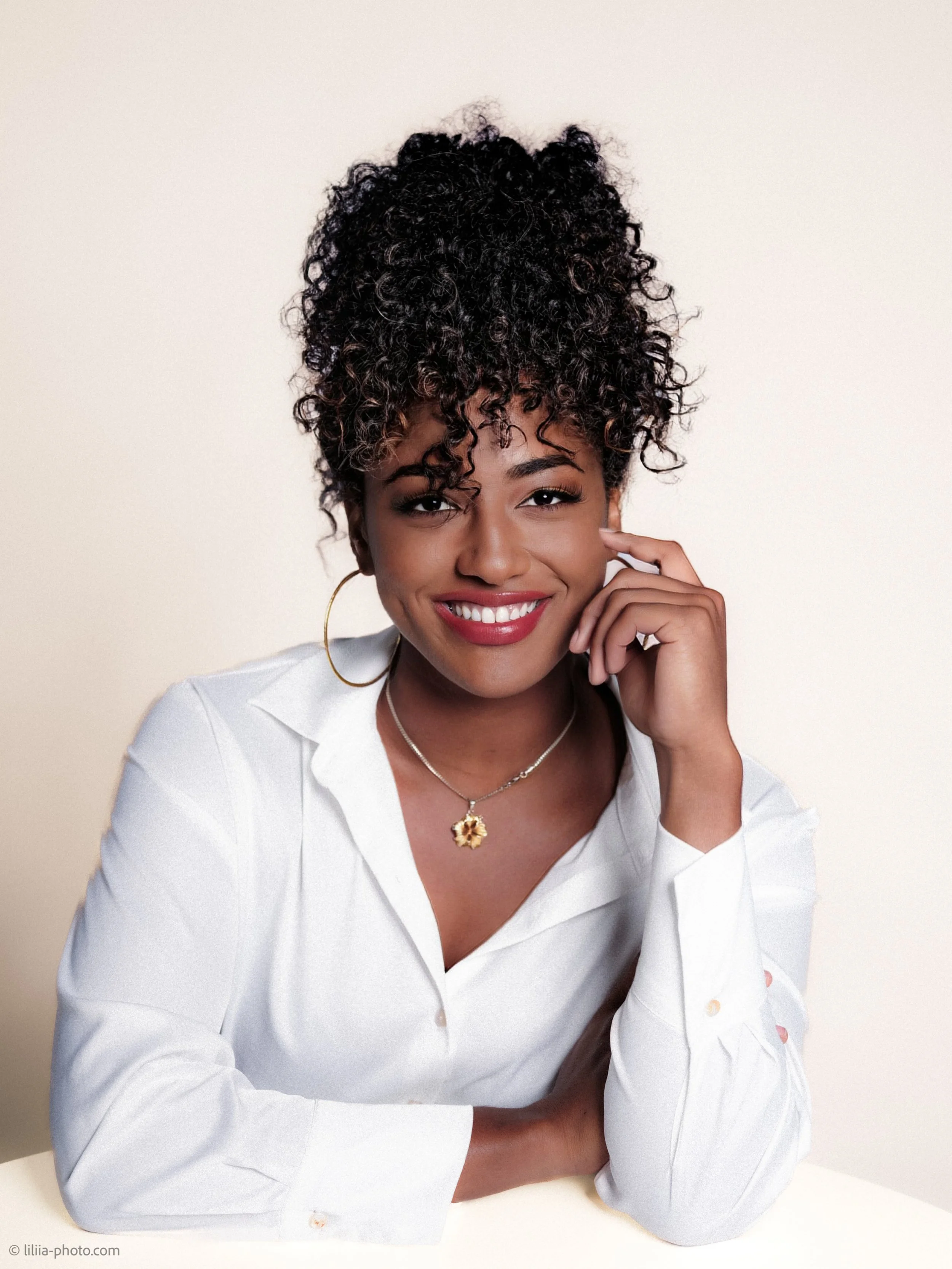 A smiling woman with curly hair wearing a white shirt and gold jewelry, resting her chin on her hand against a plain background.