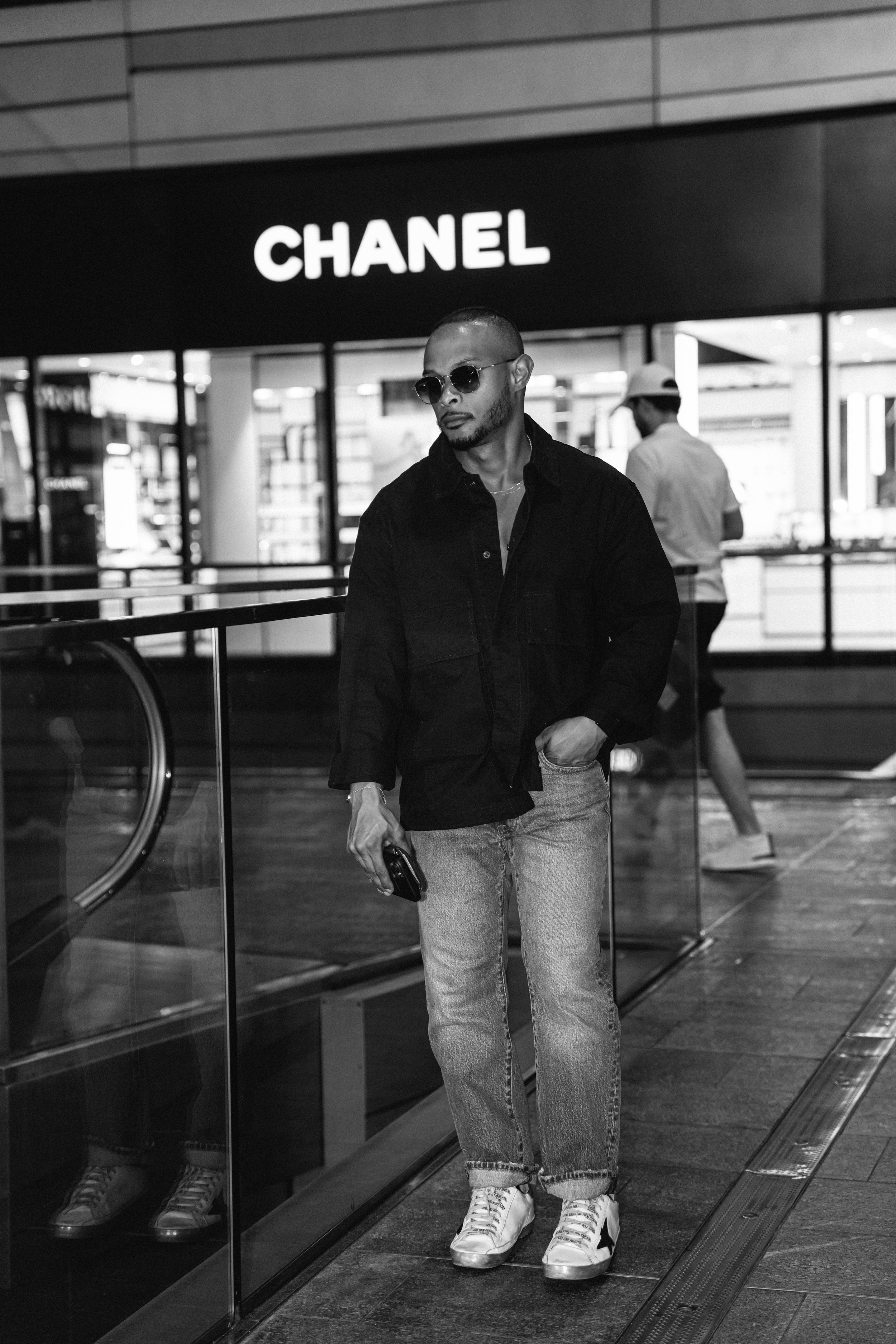 A man wearing sunglasses, a black jacket, and jeans standing inside a shopping mall near a Chanel store, holding a phone in his hand.