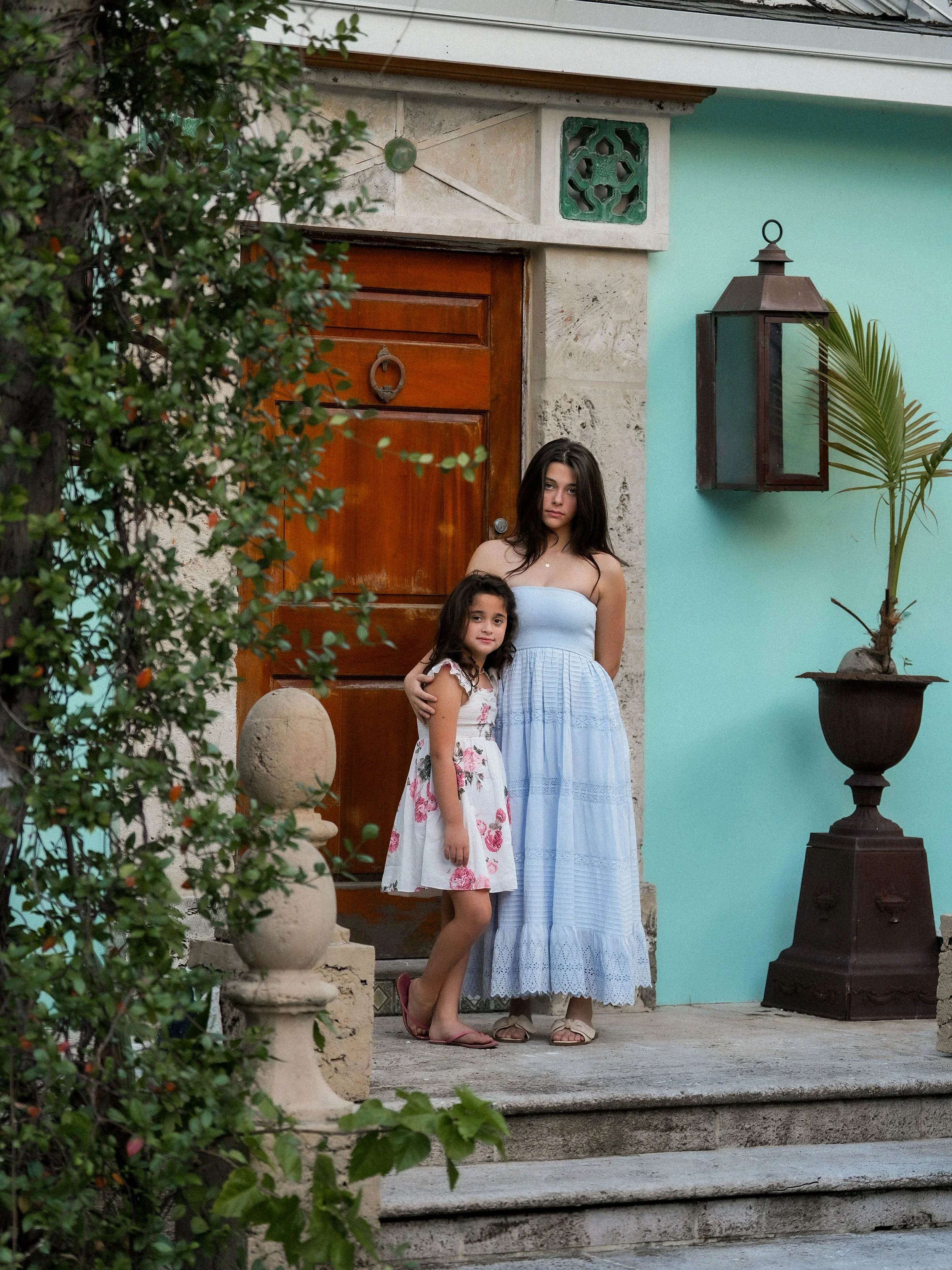 A woman and a girl standing on the front steps of a house, with the woman hugging the girl. The house has a wooden door, decorative wall features, a lantern, and a potted plant. They are both wearing summer dresses and sandals.