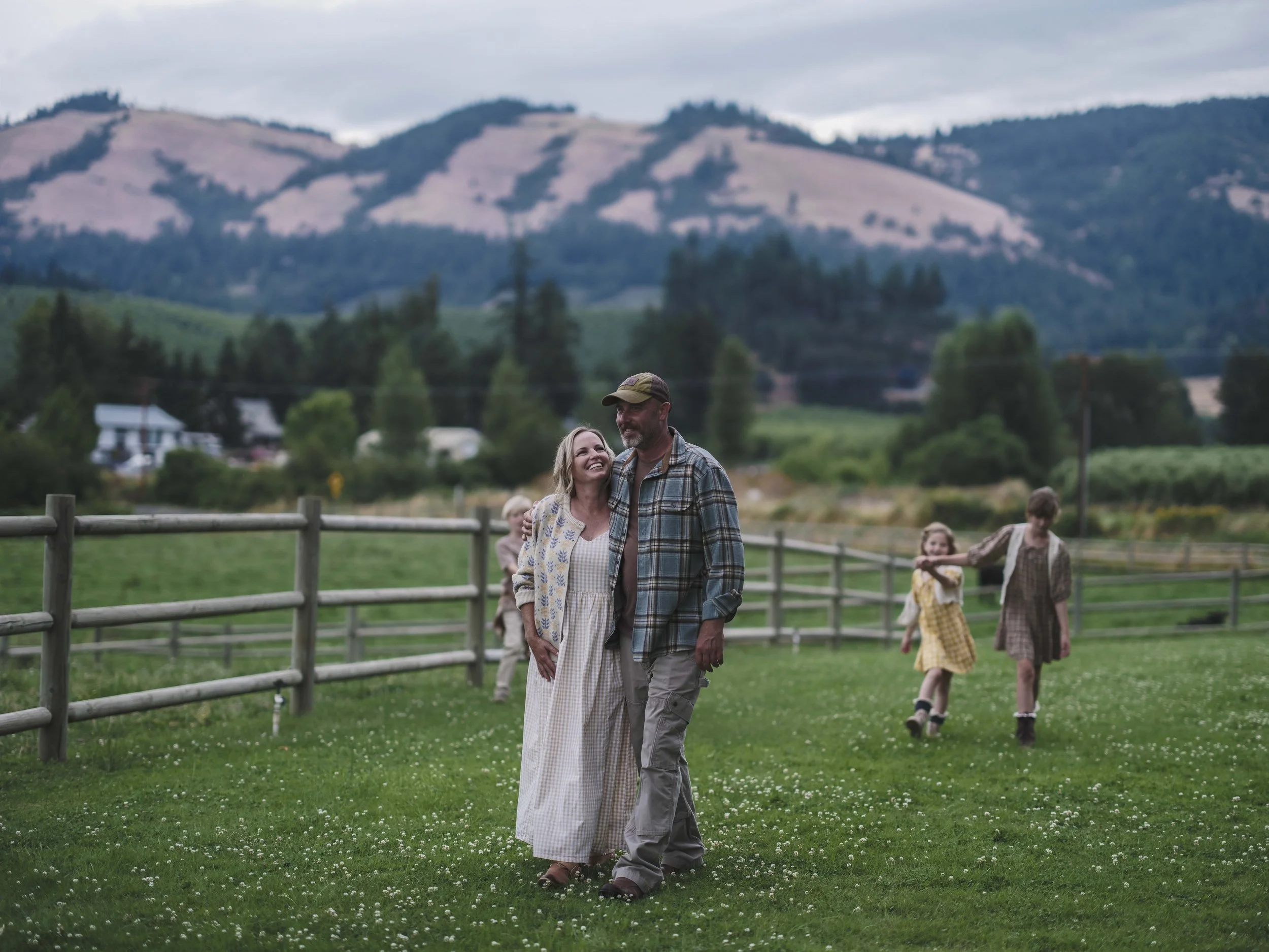 A couple walking on a grassy field with white flowers, with children playing in the background, and mountains and trees in the distance.