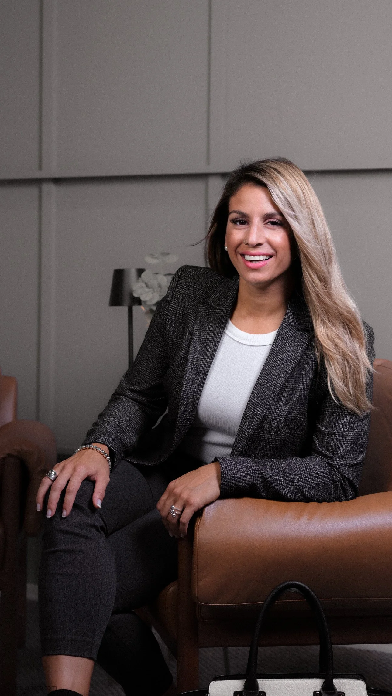 A woman sitting on a tan leather sofa, smiling and looking at the camera, in a modern office setting with a dark lamp and white flowers in the background.