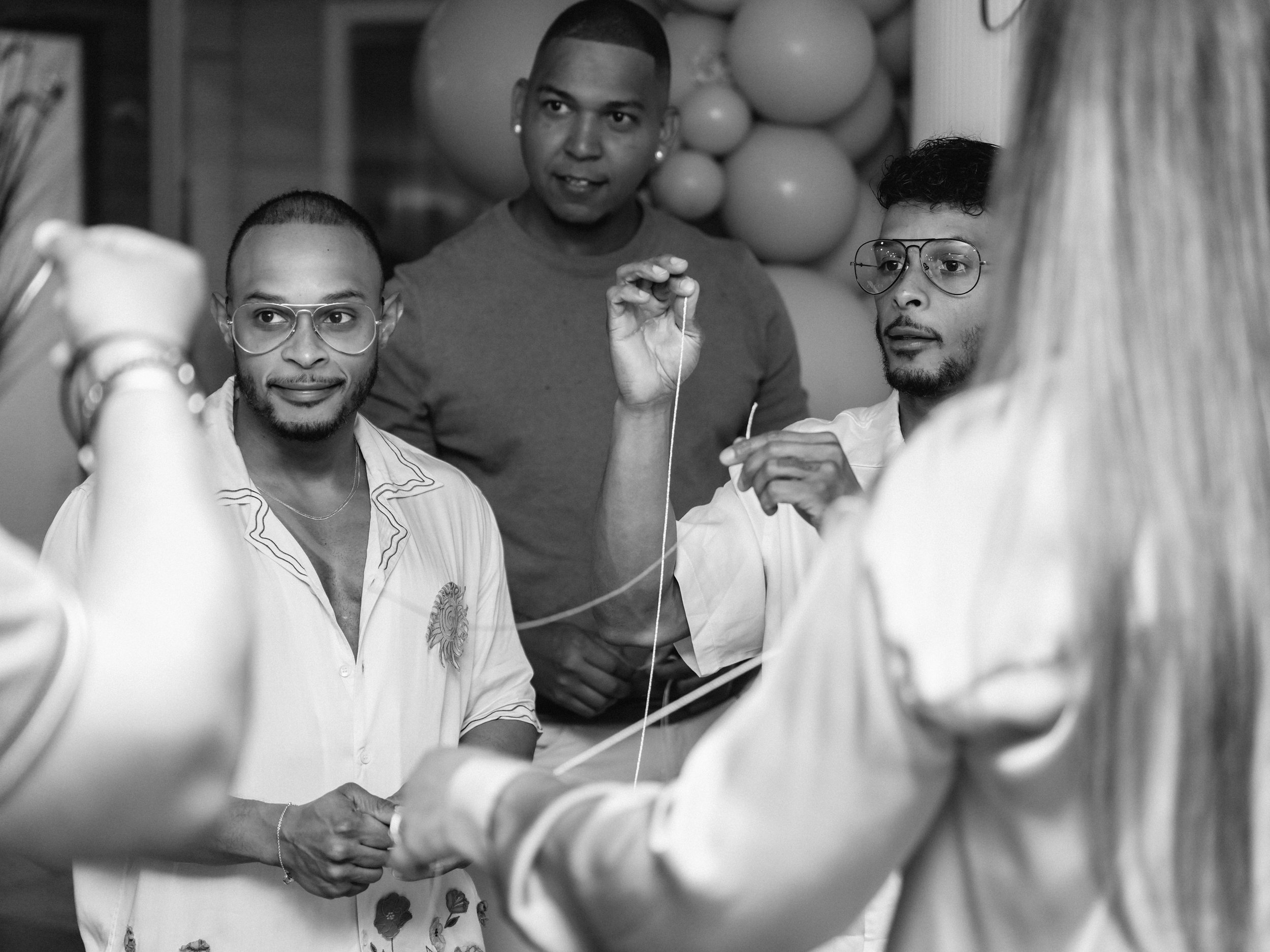 A group of men engaged in a conversation, with one man holding a string or thread, in a setting decorated with balloons.