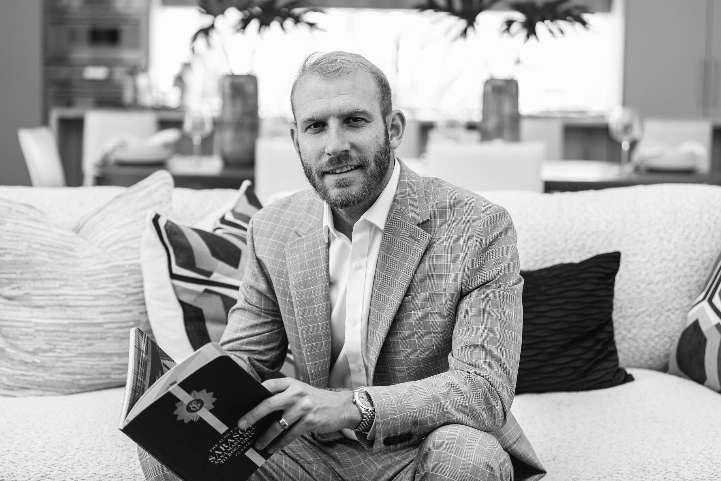 A man with a beard in a checkered suit and watch, sitting on a couch with patterned pillows, holding a book, in a well-lit room.