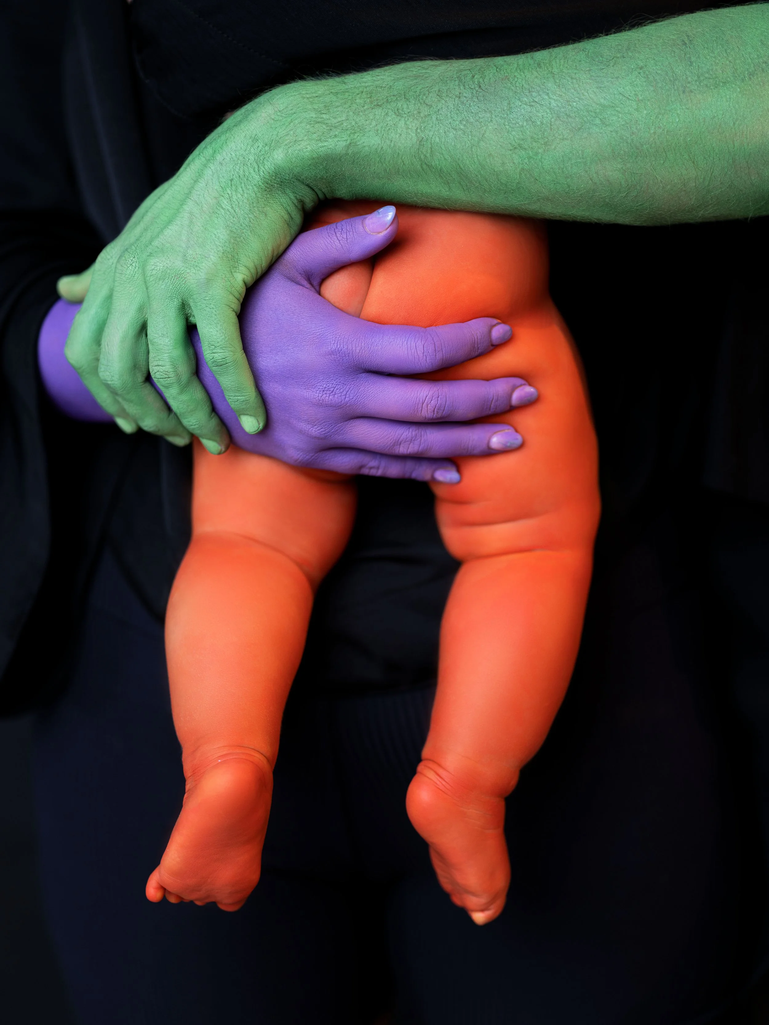 A person with one hand painted green and the other hand painted purple, holding a baby with orange painted skin.
