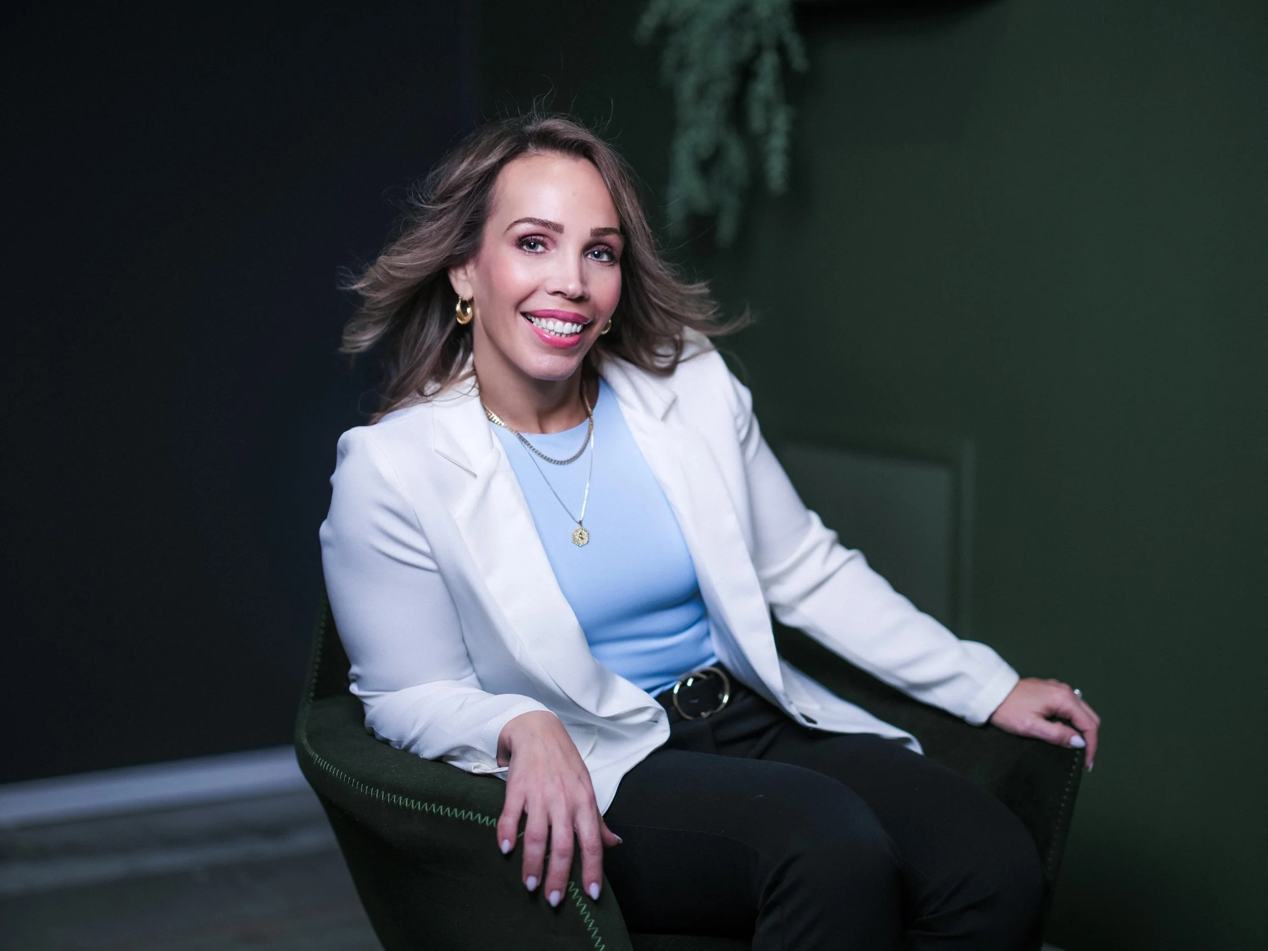A woman with wavy blonde hair smiling and sitting in a dark room, wearing a light blue blouse, white blazer, gold jewelry, and black pants.