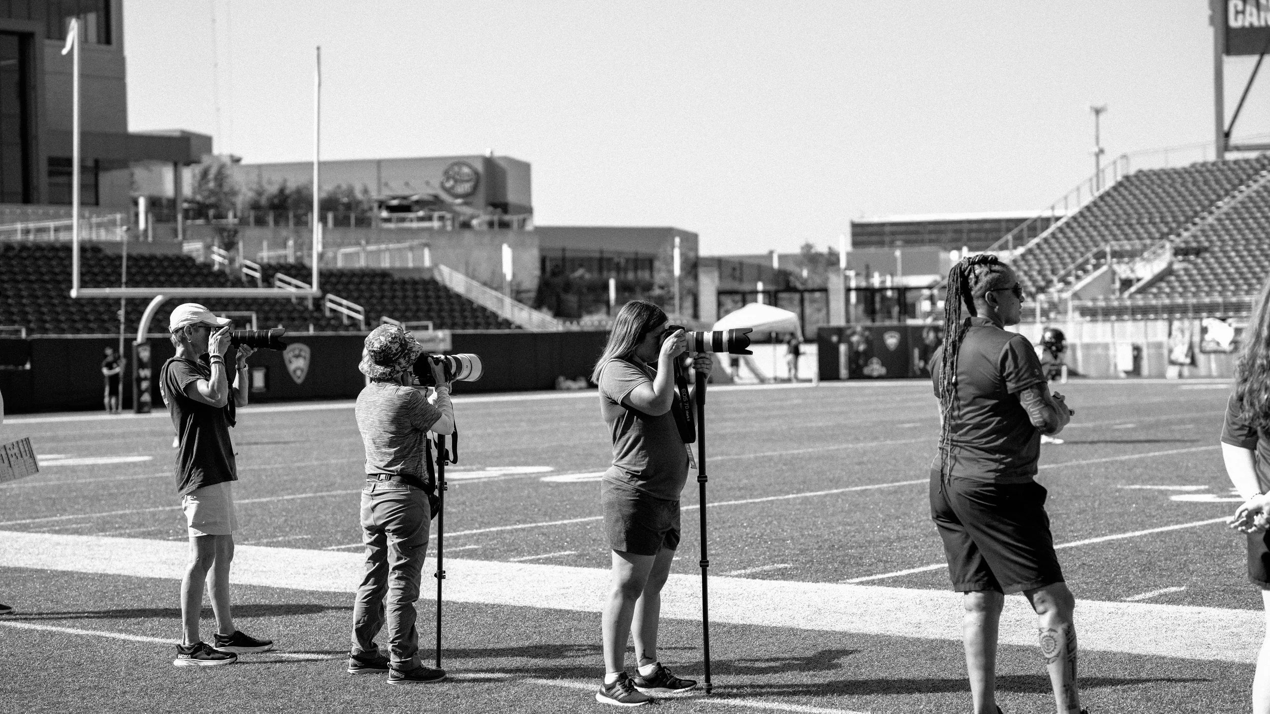Group of photographers on a football field, taking pictures with professional cameras with long lenses, during daytime in a stadium.
