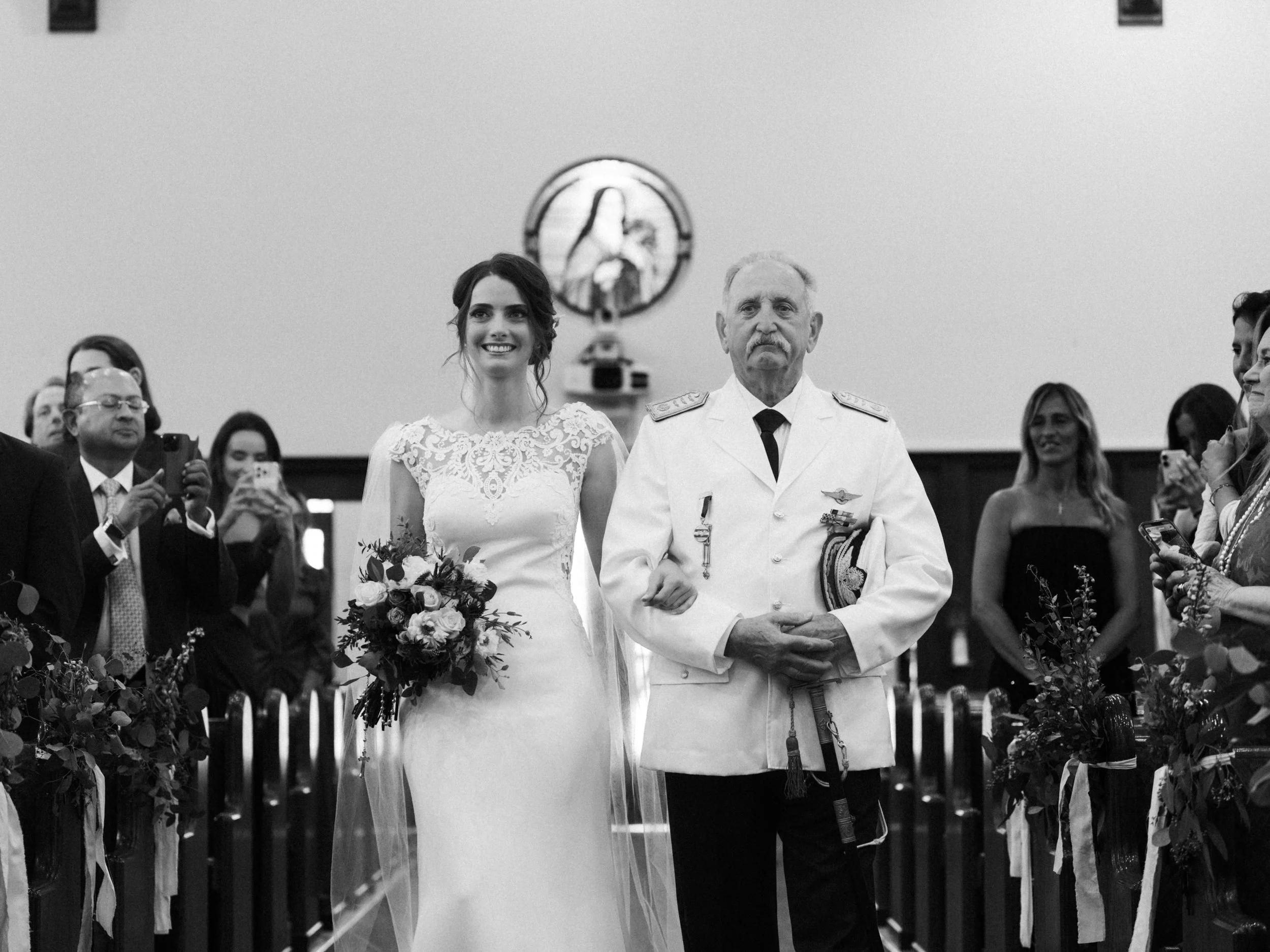 A bride in a wedding dress walking down the aisle accompanied by a man in a military uniform, surrounded by guests taking photos in a church.
