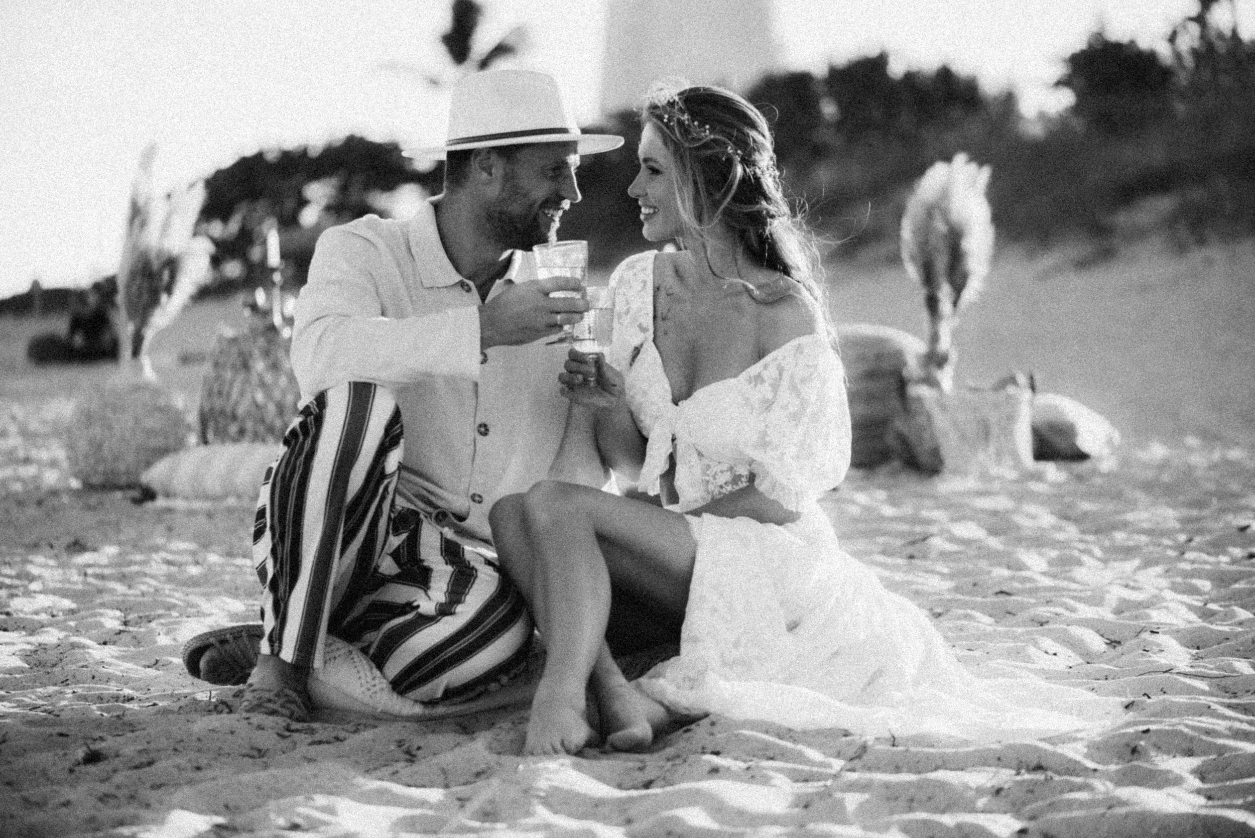 A man and woman sitting on the sandy beach, smiling at each other and holding drinks, with the woman wearing a white lace dress and the man wearing striped pants and a light-colored shirt and hat.