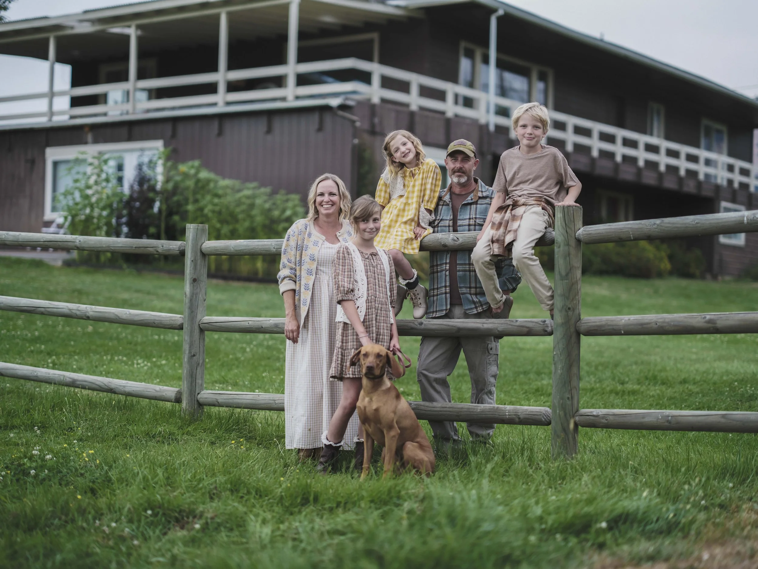 Family of six with a dog standing outside on a grassy area in front of a wooden fence, with a house in the background.
