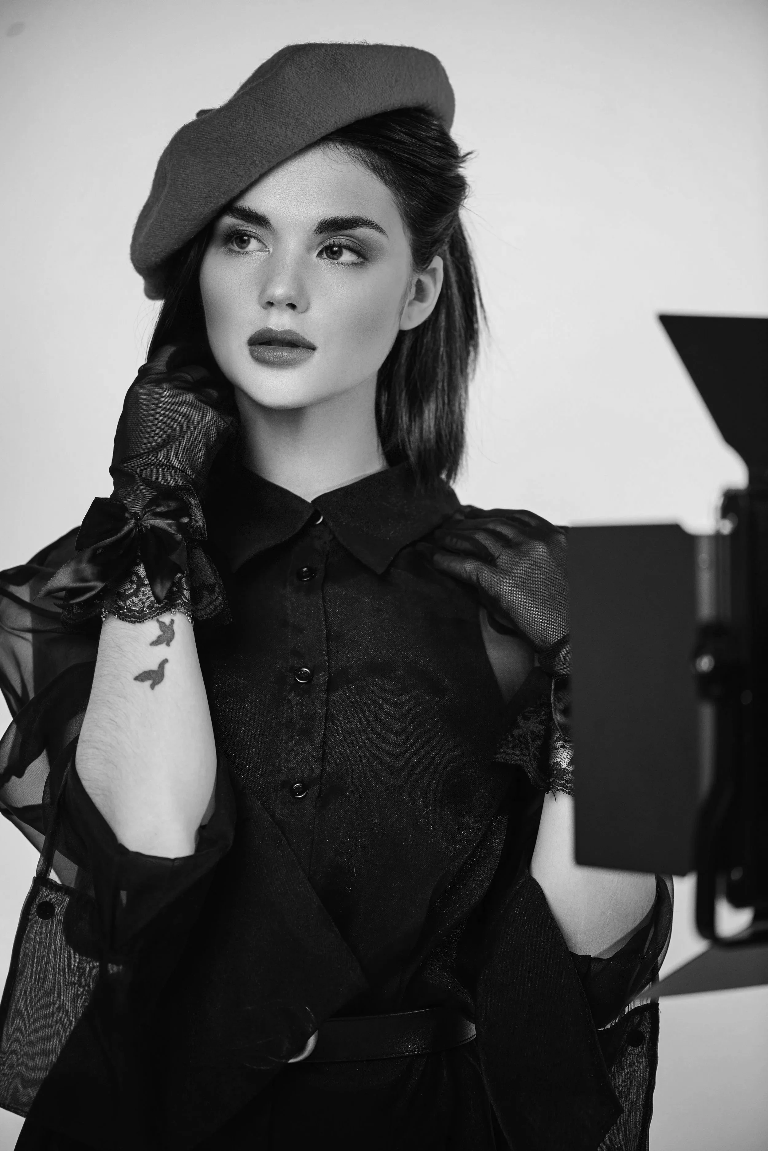 A black and white photo of a woman wearing a beret, gloves, and a dark outfit with lace details, posing in front of a camera.