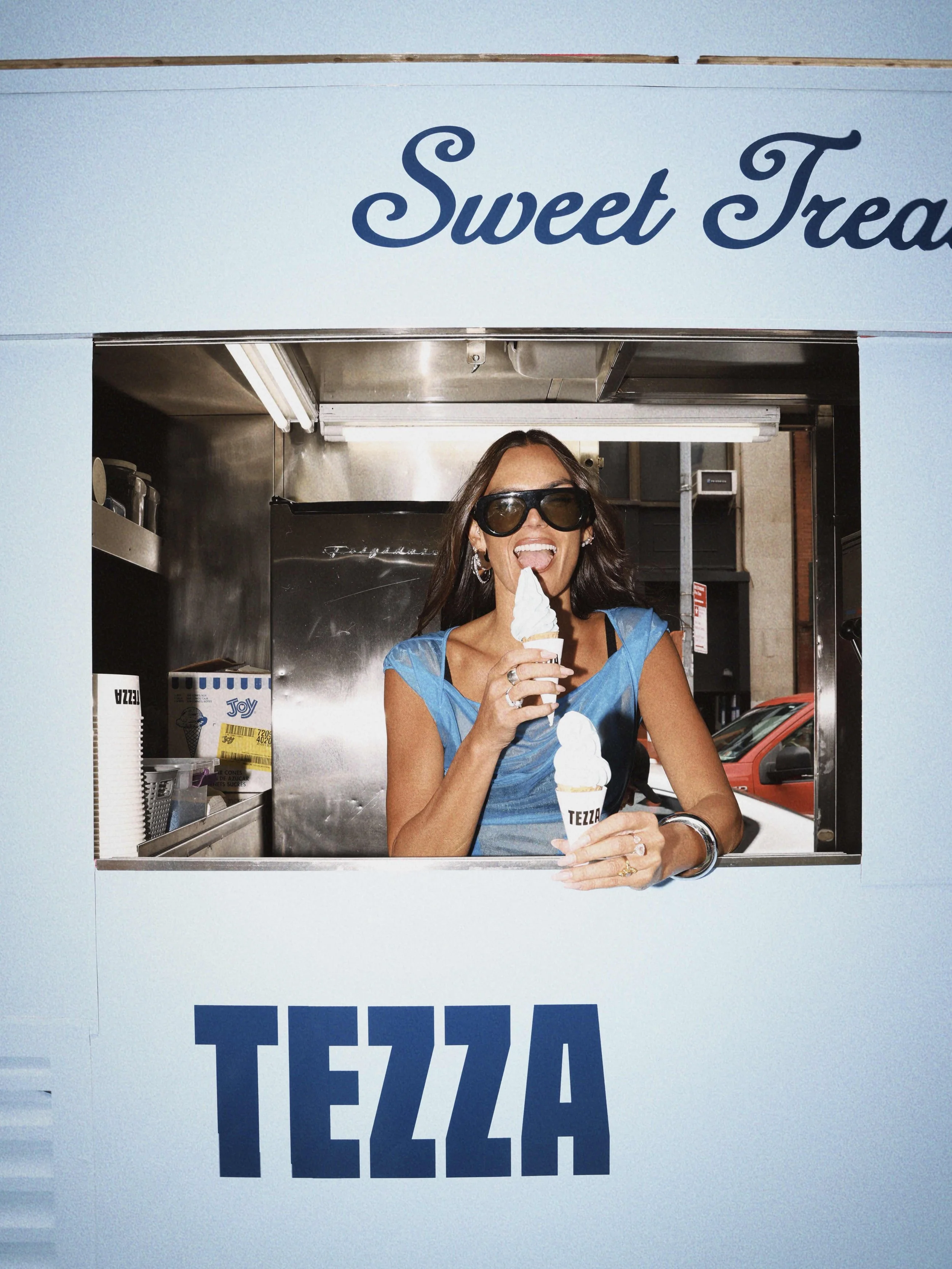 A woman with long dark hair and sunglasses enjoying ice cream in a pastel blue ice cream truck with the words 'Sweet Treats' and 'TEZZA' written on it.