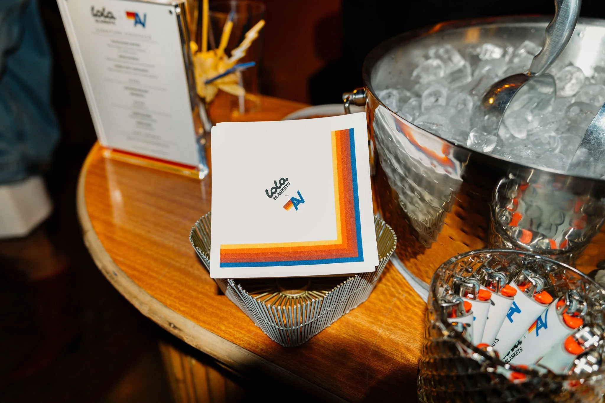 A table with a stack of white napkins bearing the logo 'Lalo Blankets' and a multicolored stripe, a metal ice bucket filled with ice and a serving spoon, and a container with individually wrapped orange and white lollipops.