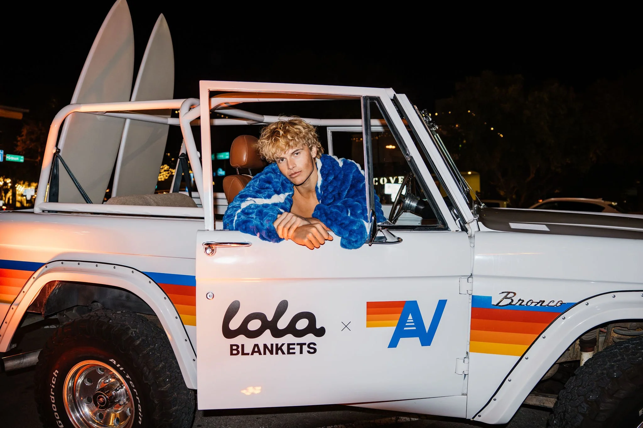 A person with curly blonde hair wearing a blue fuzzy jacket sitting in a white vintage Ford Bronco parked at night, with surfboards attached to the back and a cityscape with lights in the background.