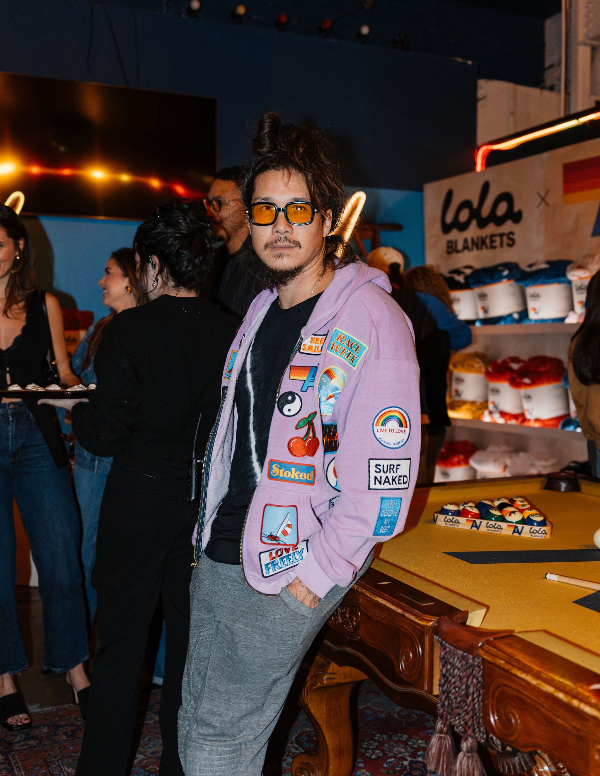 A man with glasses and a mustache wearing a lavender jacket with patches, standing at a social gathering.
