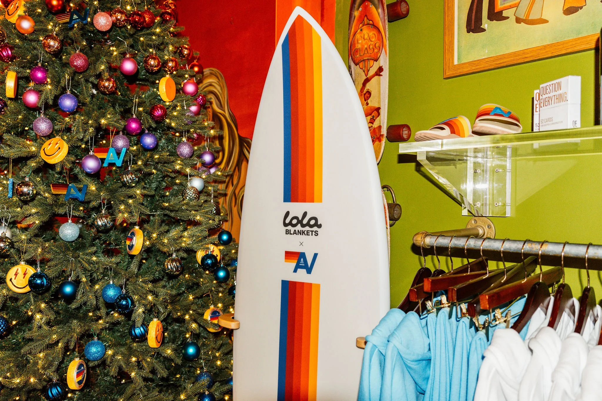 Interior of a store decorated for Christmas with a Christmas tree adorned with pink, purple, blue, silver, and gold ornaments, some with smiley face and rainbow designs. A surfboard with colorful stripes and the text "Lola Blankets" and "N" is positi