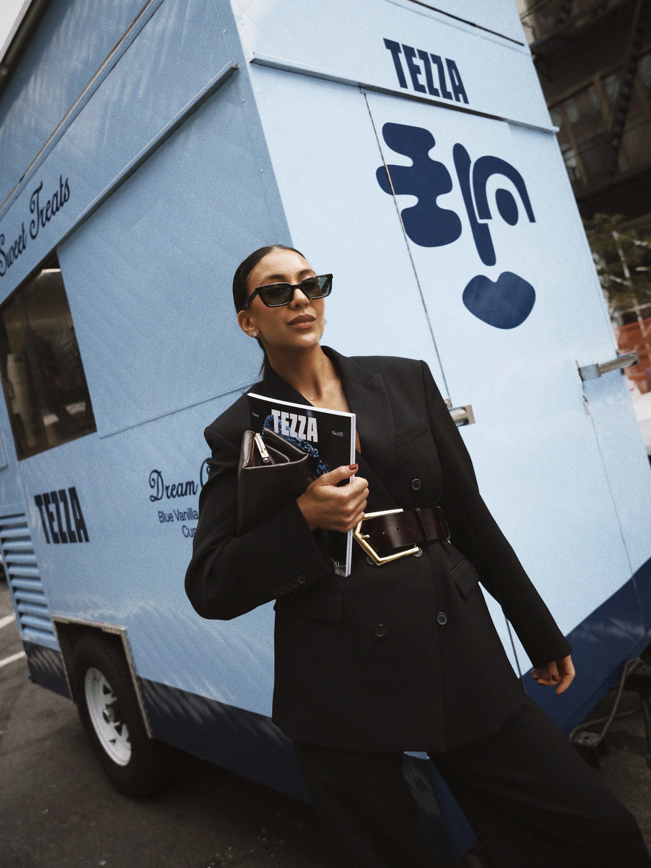 A woman in a black blazer and pants, wearing sunglasses and holding a brochure, stands next to a blue ice cream truck with the word 'TEZZA' and a heart logo.