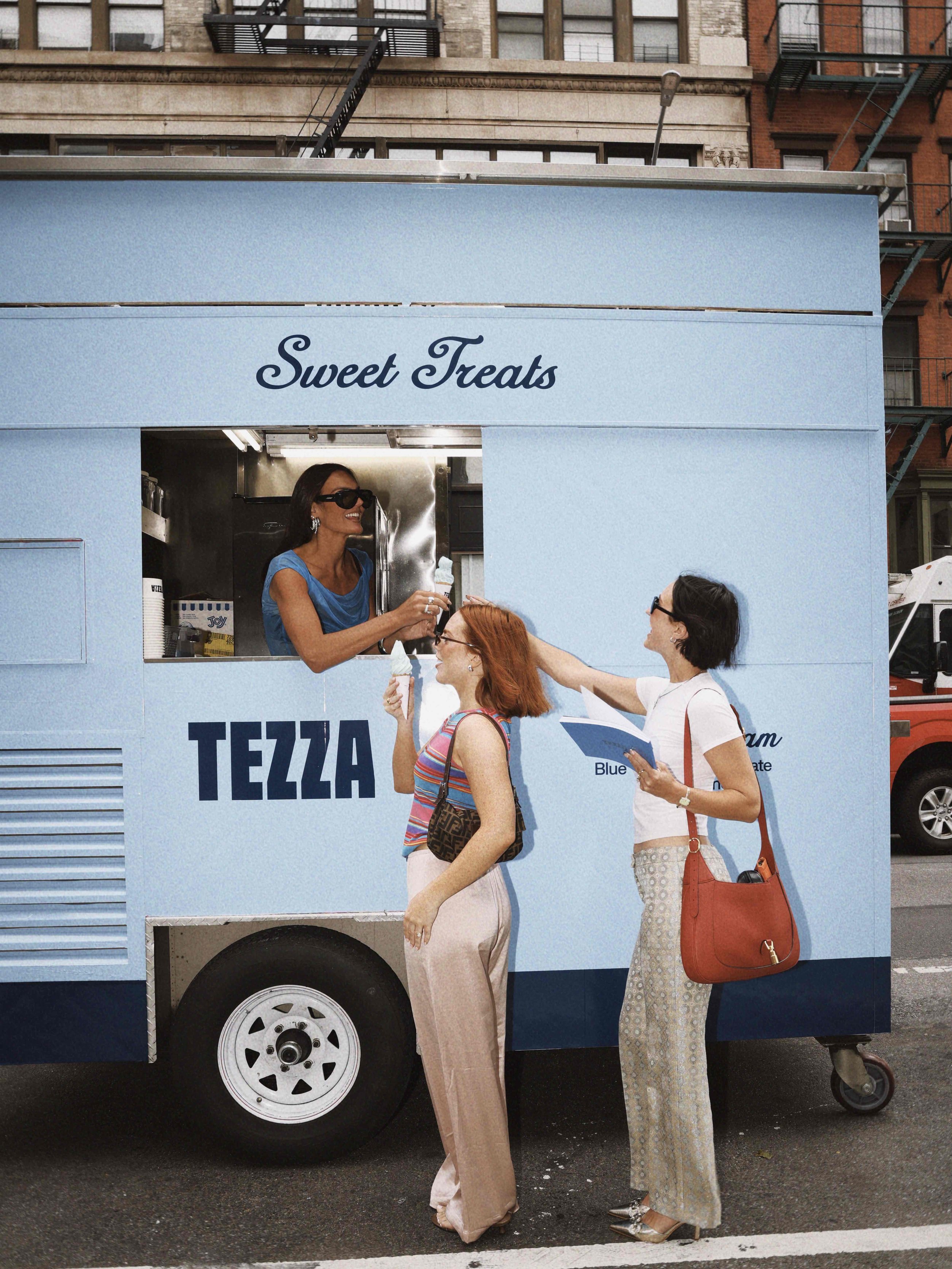 Three women at a blue ice cream truck called 'Sweet Treats,' two of them are receiving ice cream from the woman inside the truck. One woman is holding an ice cream cone, and another woman is holding a menu or brochure.
