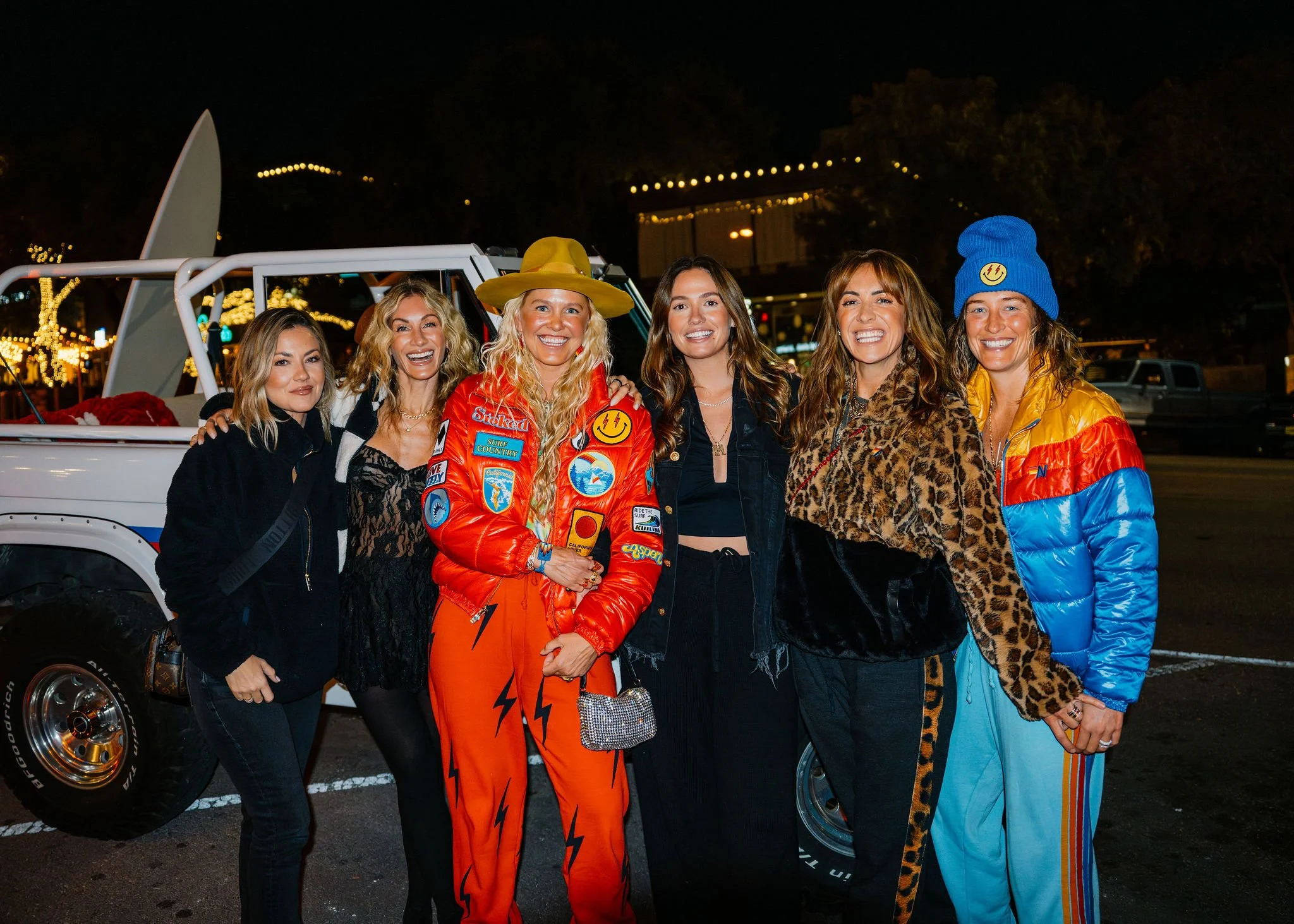 Six women smiling at night, standing in front of a white vehicle with gambling chips and a red blanket inside. One woman is wearing a bright red jacket with patches, an orange hat, and holding a small patterned purse. The others are dressed in variou