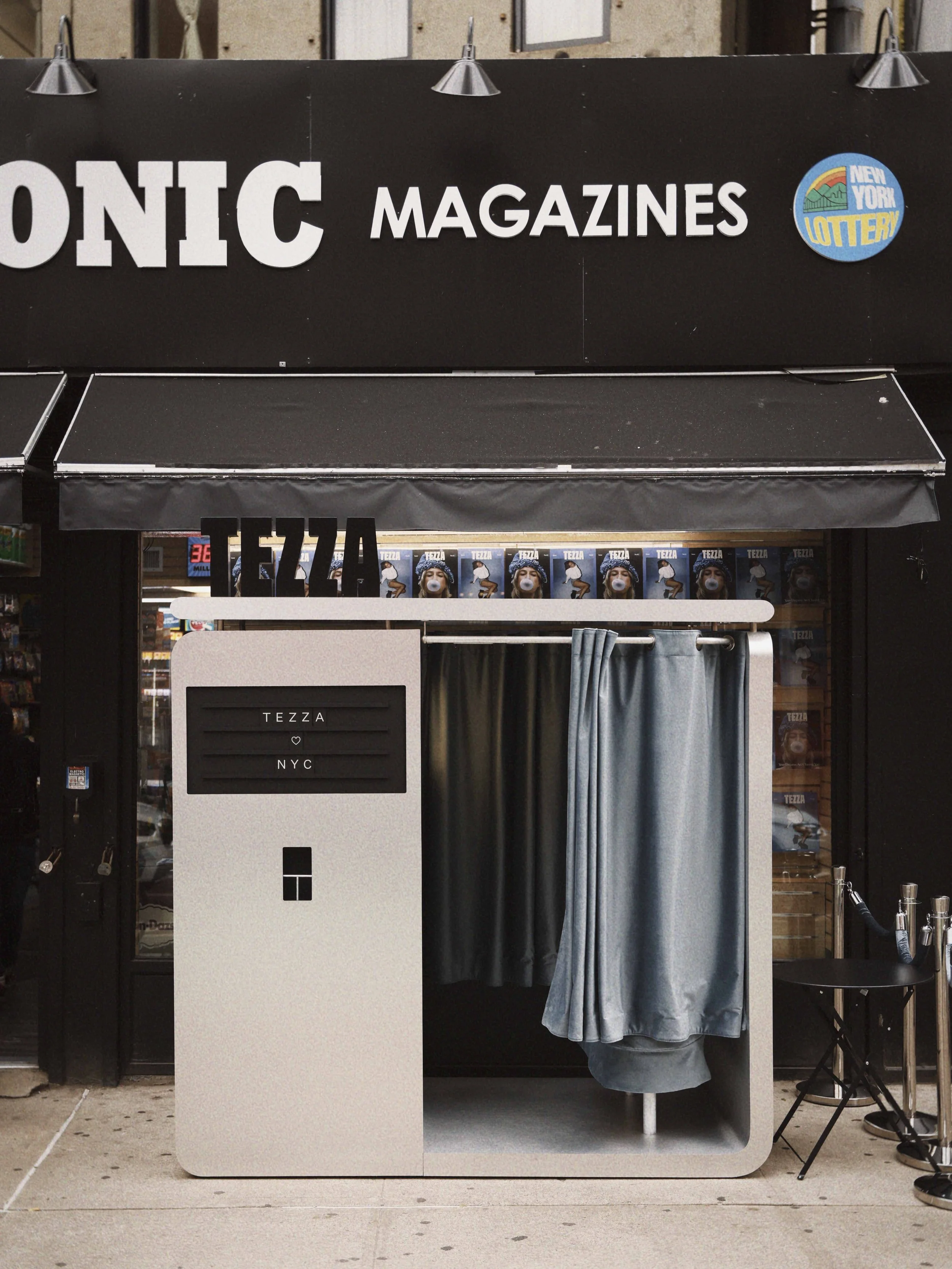 A photo of a photo booth outside a magazine store with a sign that reads 'TEZZA NYC'. The booth has grey curtains and a sign with 'TEZZA'.