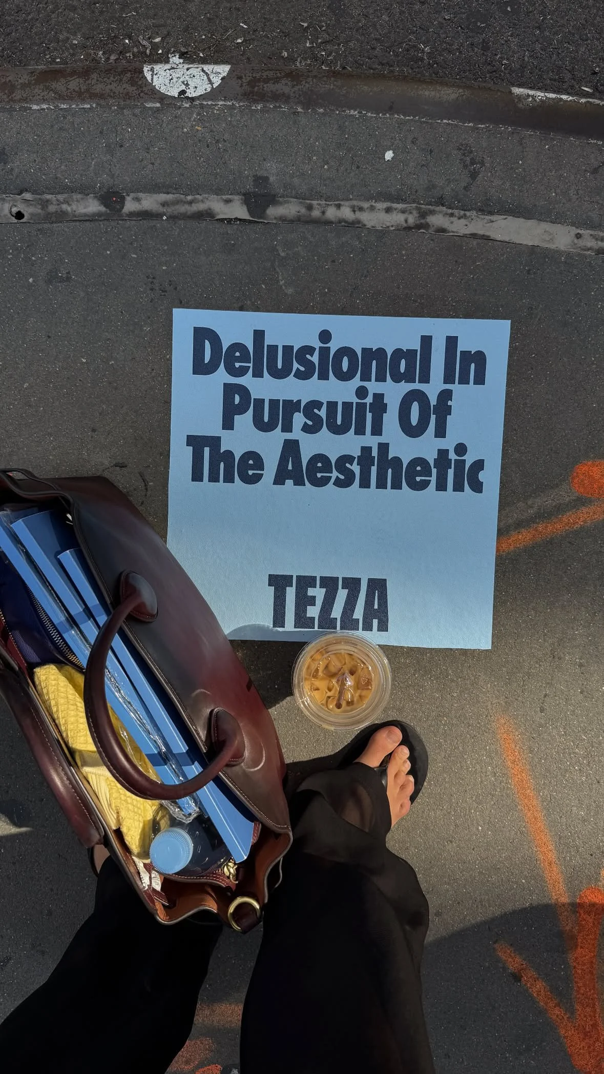 A poster on the ground reads "Delusional In Pursuit Of The Aesthetic TEZZA" next to a person's foot, a bag with various items, and a cup of iced coffee, on a city street.