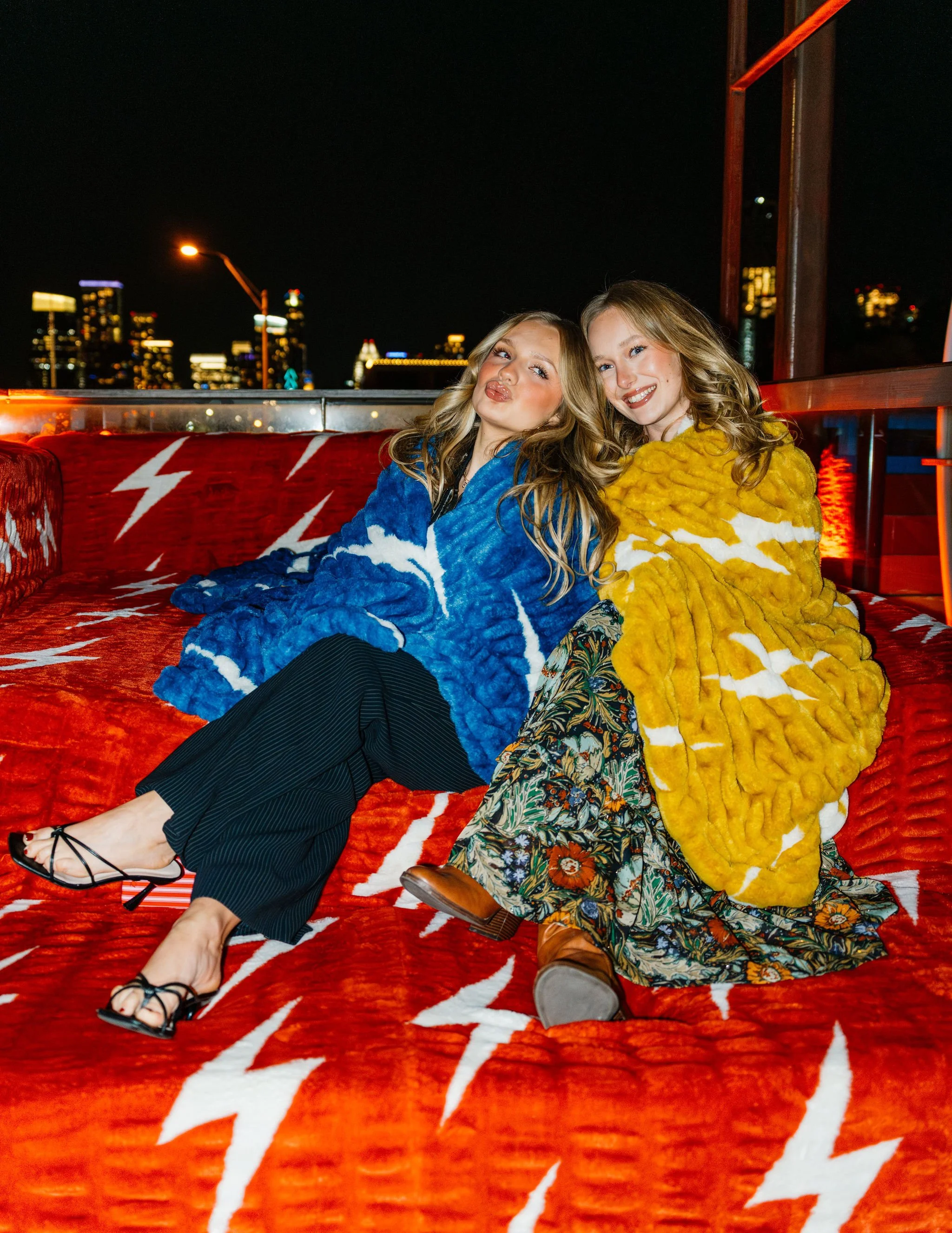 Two young women sitting on a red, lightning bolt patterned couch at night with city skyline in the background. One woman wears a blue textured coat, and the other wears a yellow textured coat, both smiling and posing for the camera.