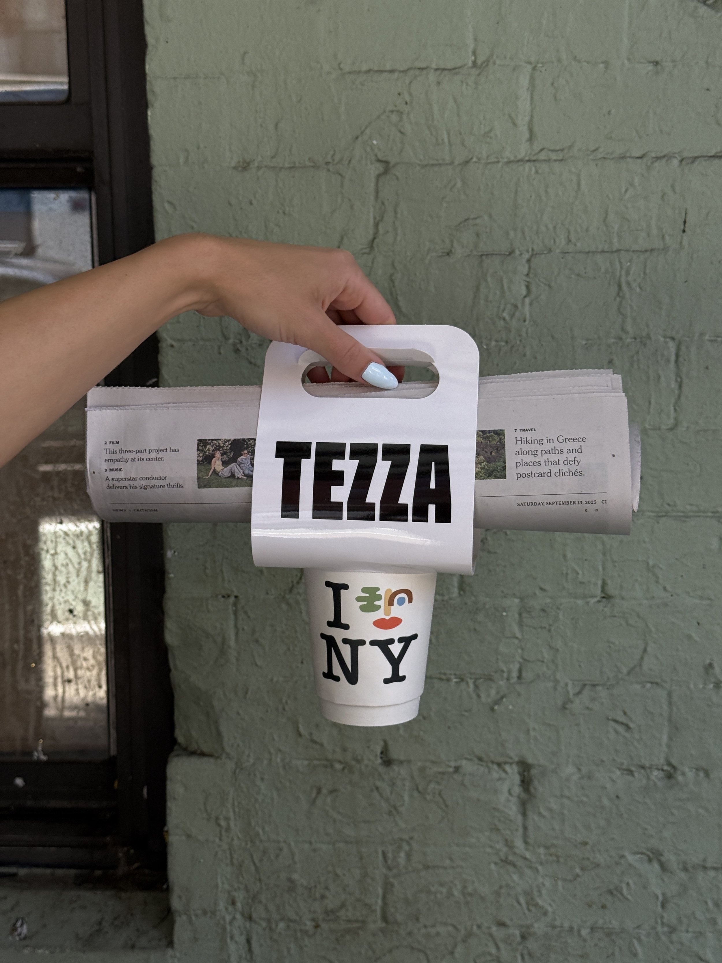 A hand holding a coffee cup with 'I ❤️ NY' design, with a newspaper rolled into a tubular shape that has 'TEZA' written on a label attached to it.