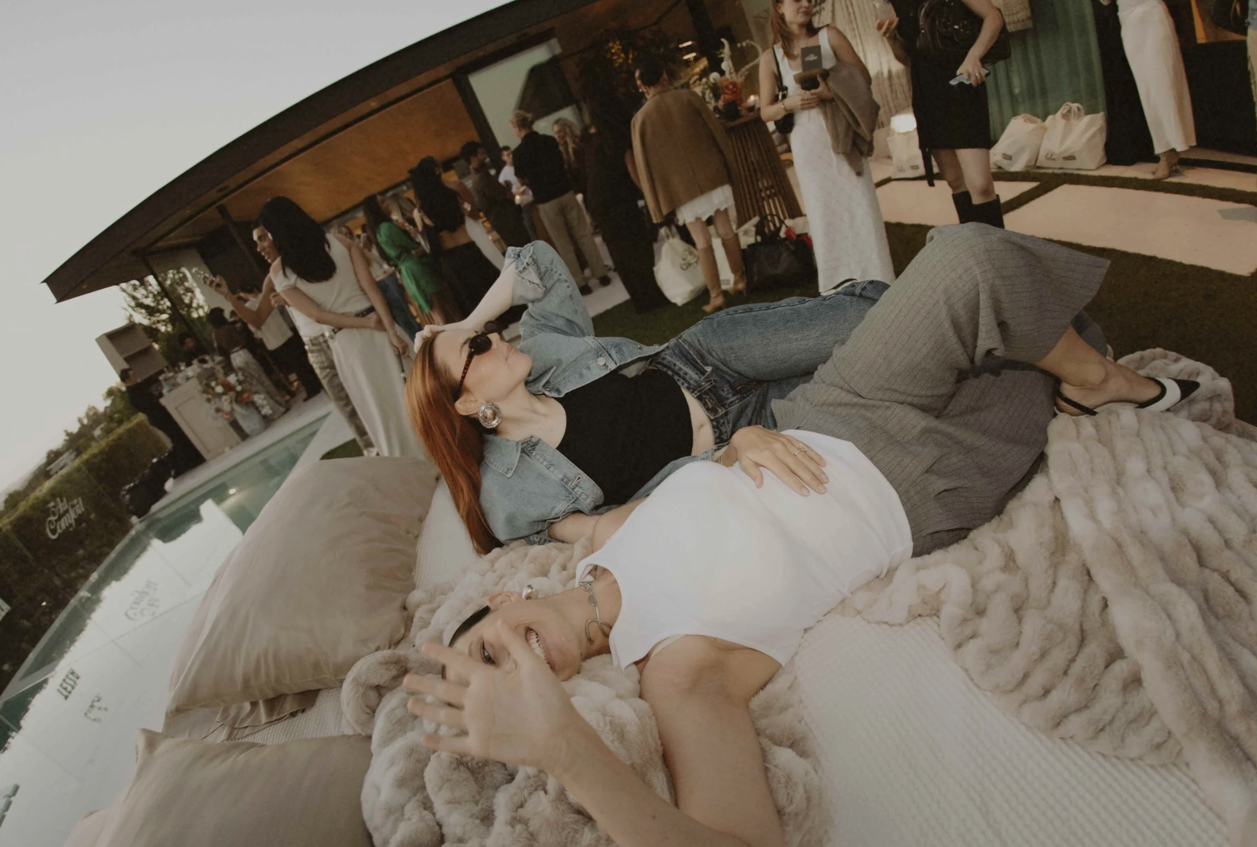 Two women are lying on a bed outdoors at a social gathering, with people standing and chatting in the background.