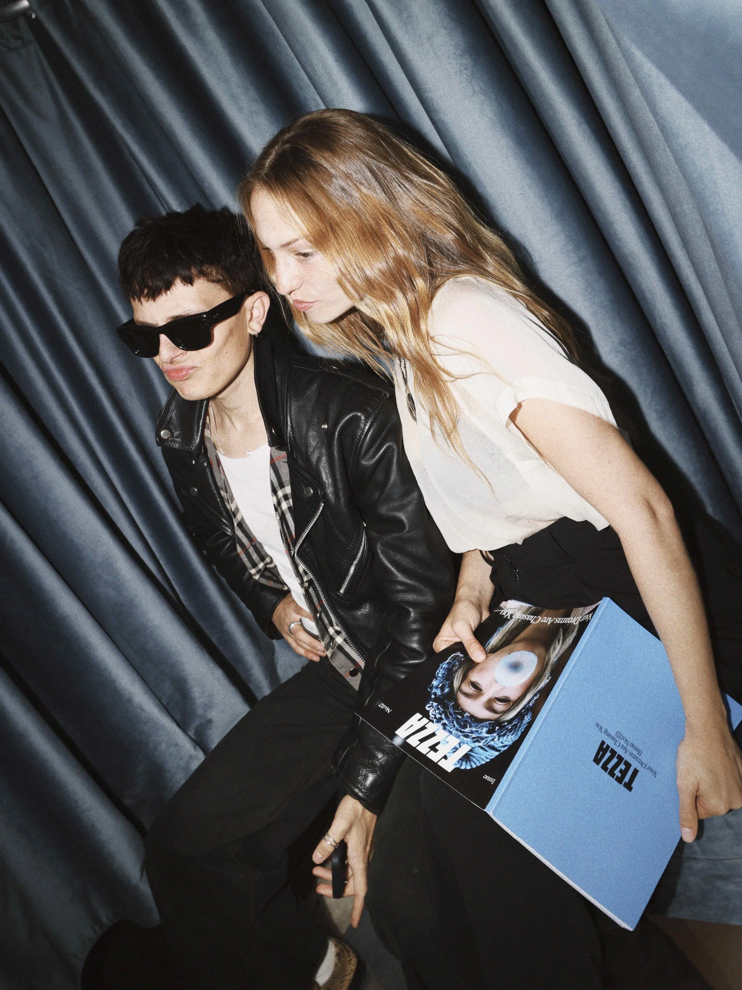 Two young adults, one with short dark hair wearing sunglasses and a leather jacket, and the other with long red hair in a white top, sitting against a backdrop of dark blue curtains. The person with red hair holds a large blue box featuring a photo o