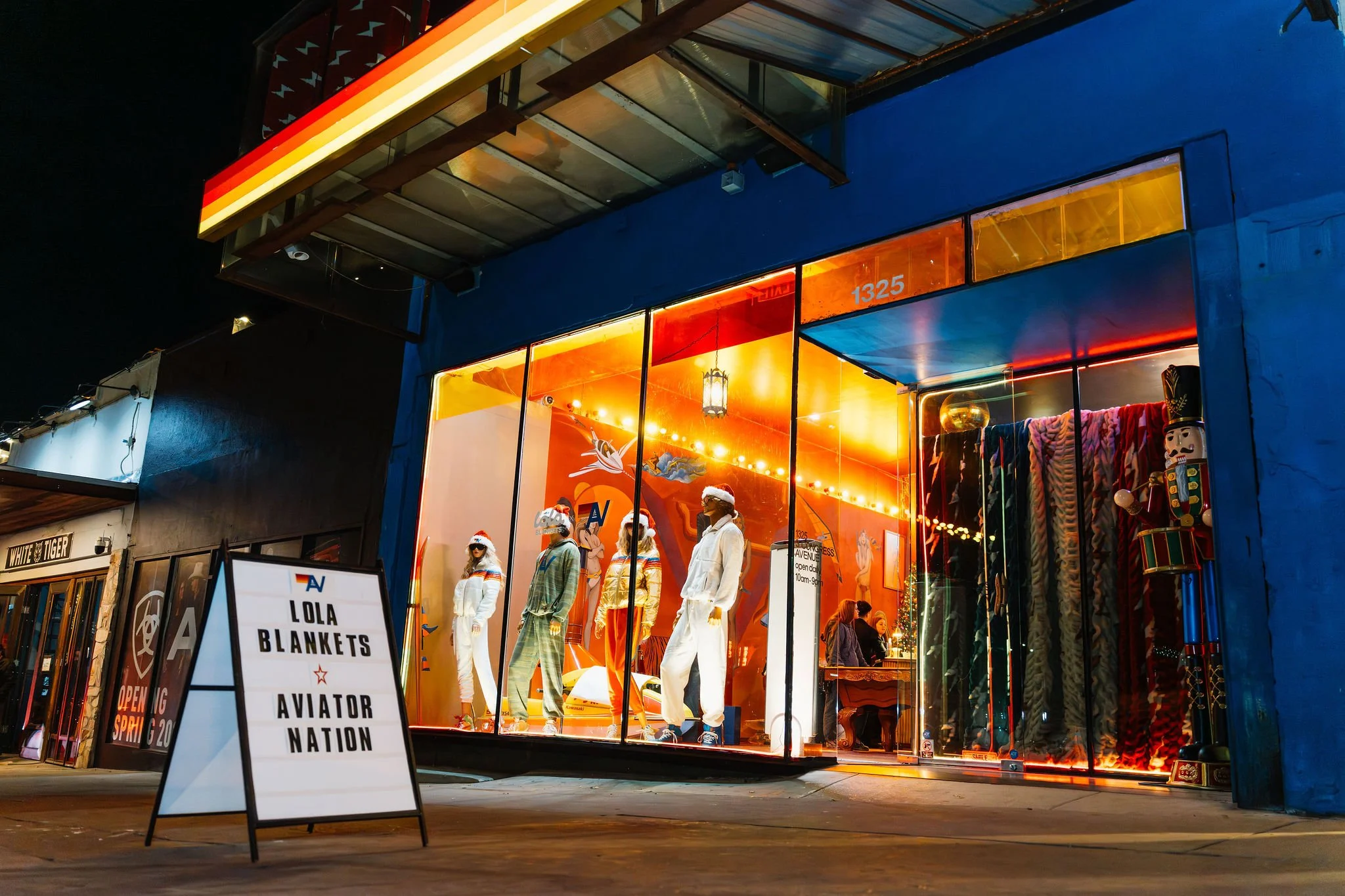 Nighttime view of a retail store window display featuring mannequins dressed in pajamas, holiday accessories, and Santa hats. The store has bright warm lighting inside, with colorful clothing and a large toy soldier figure inside. A sign outside read