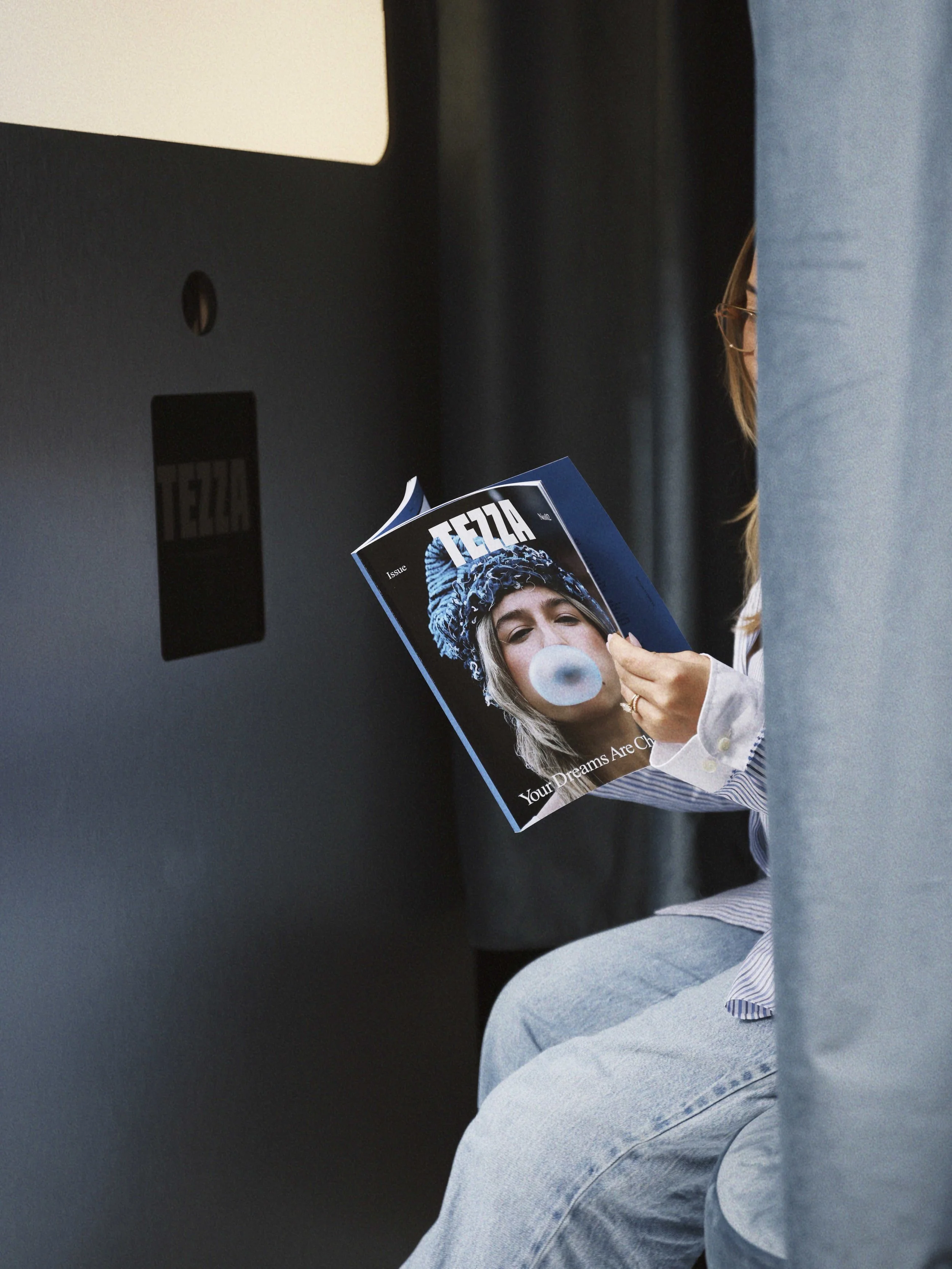 Person sitting on an airplane seat, partially hidden behind a curtain, reading a magazine with a woman blowing bubblegum on the cover.