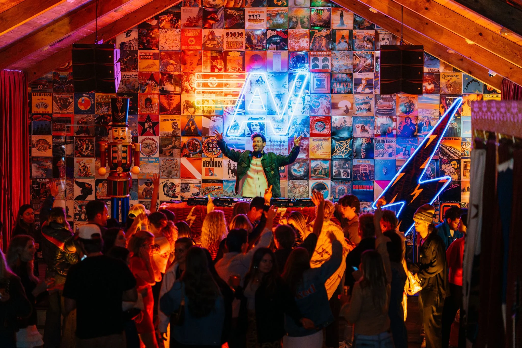A DJ performs on stage at a lively nightclub with a large crowd dancing. The stage background features a collage of colorful album covers, neon signs, including a lightning bolt and a triangle, and decorative elements. The scene is illuminated with vibrant neon and ambient lighting.