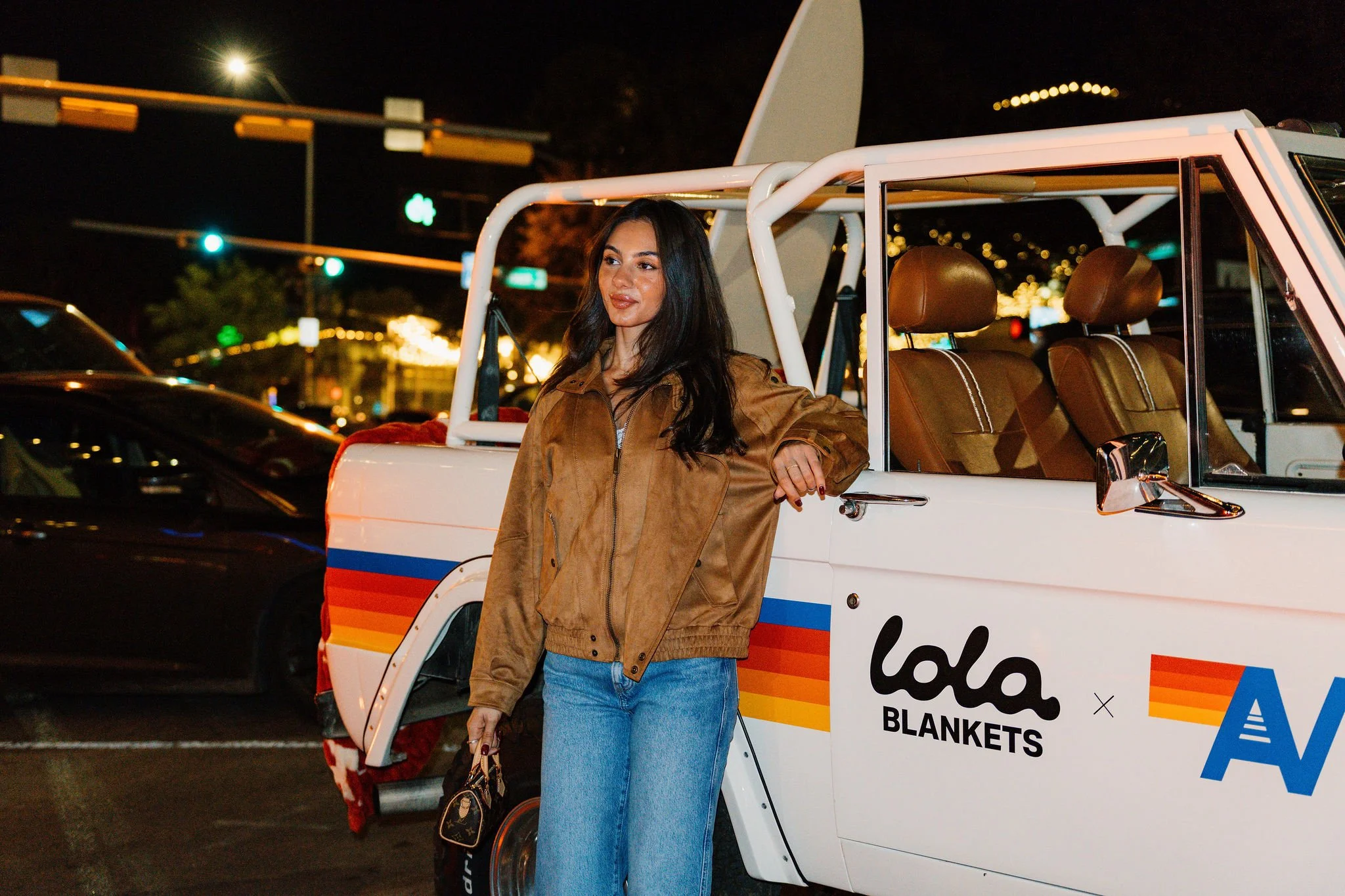 A woman with long dark hair leaning against a white vehicle with colorful stripes, at night, in an urban setting.
