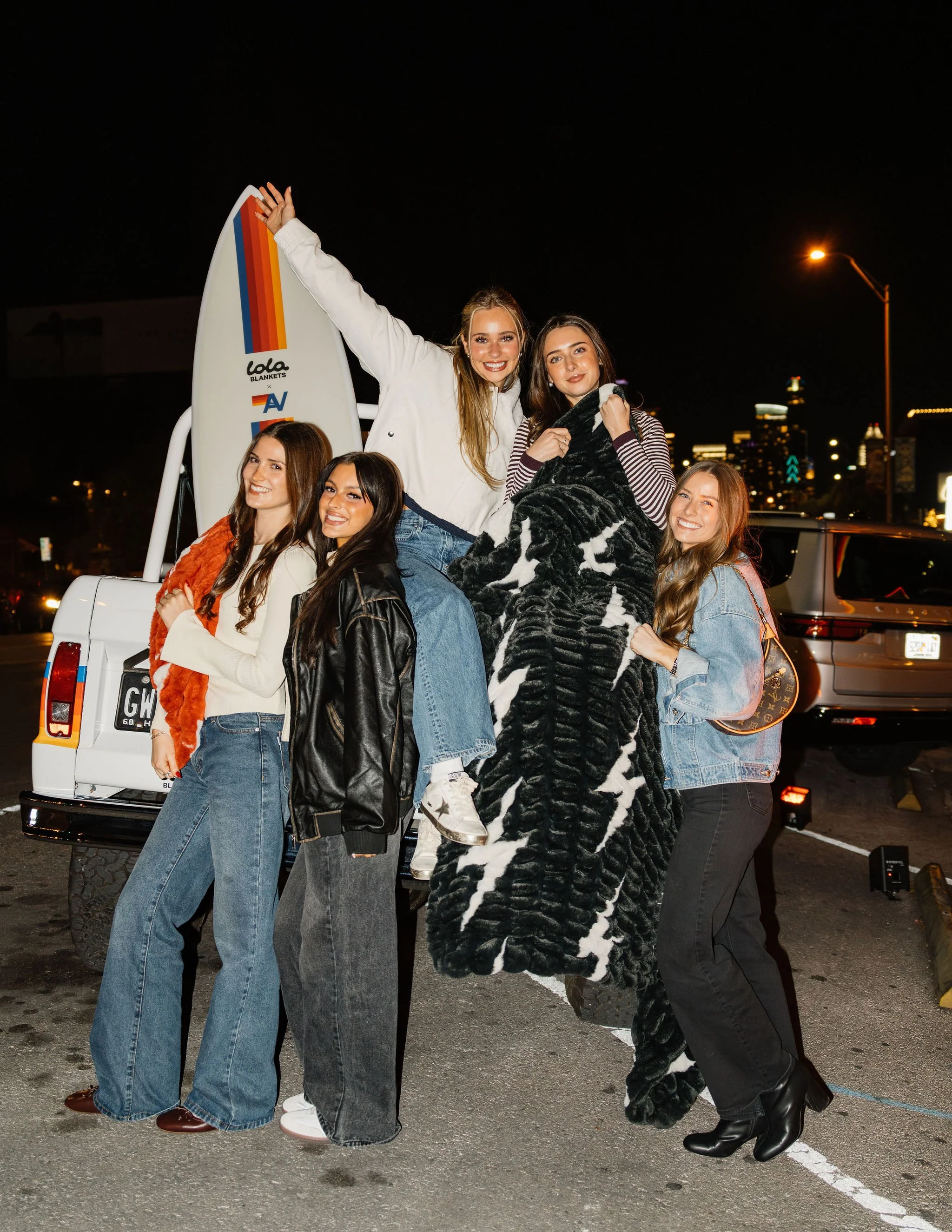 Group of six young women celebrating at night in a parking lot, with a white vehicle in the background, some holding a blanket and a surfboard.