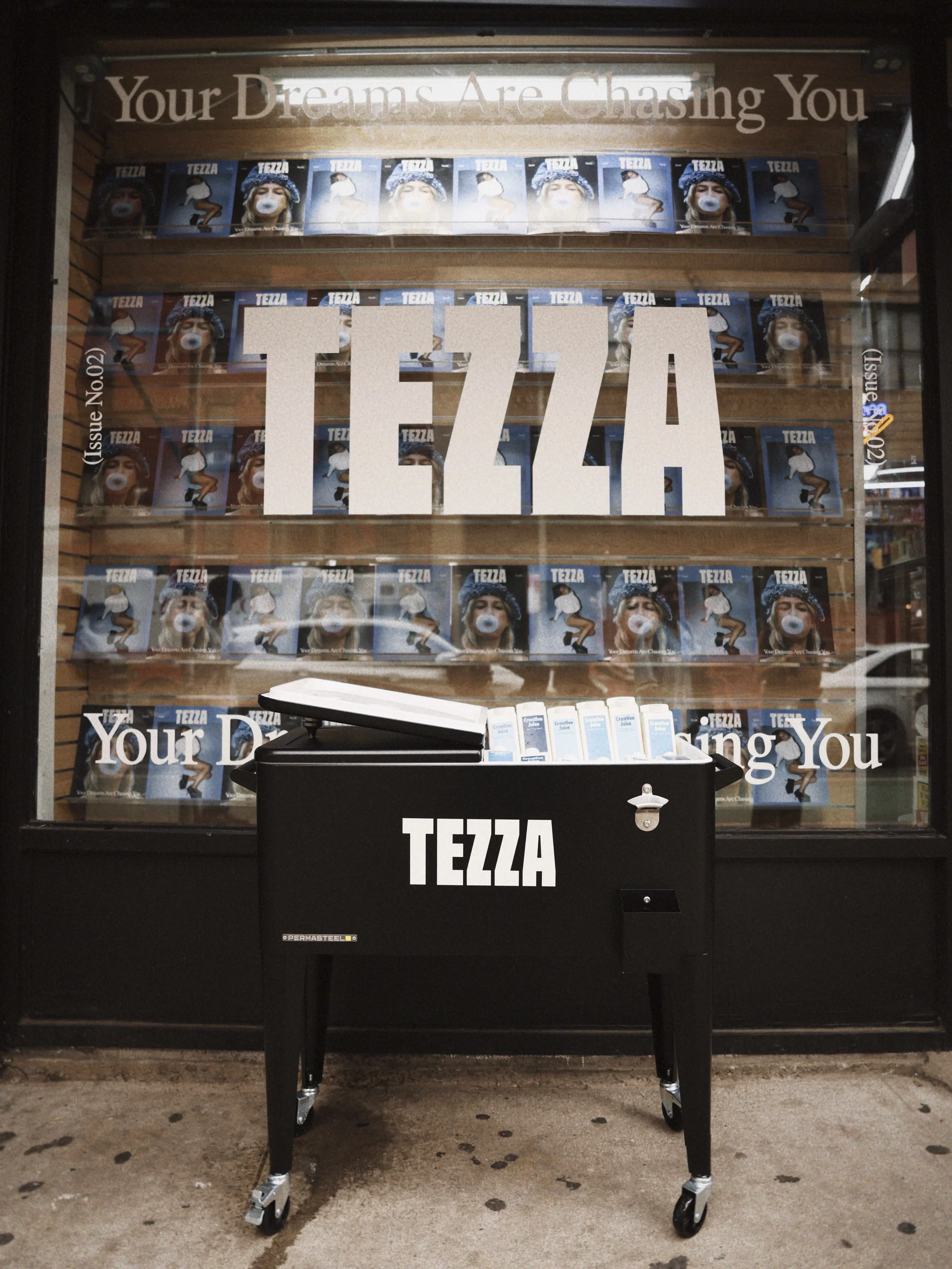 Store window display with the words 'Your Dreams Are Chasing You' and a large 'TEZZA' sign. A black cart with the word 'TEZZA' on it is in front of the window, containing copies of a magazine or book titled 'TEZZA.'