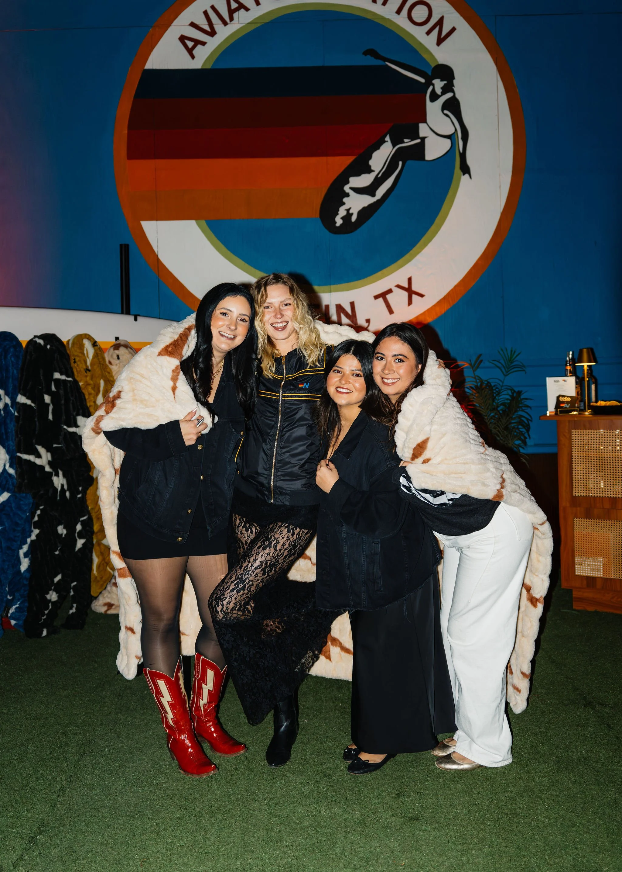 Four women smiling and posing together at an indoor event, with a large circular logo for Austin, TX, with a skier graphic in the background, and wearing warm clothing.