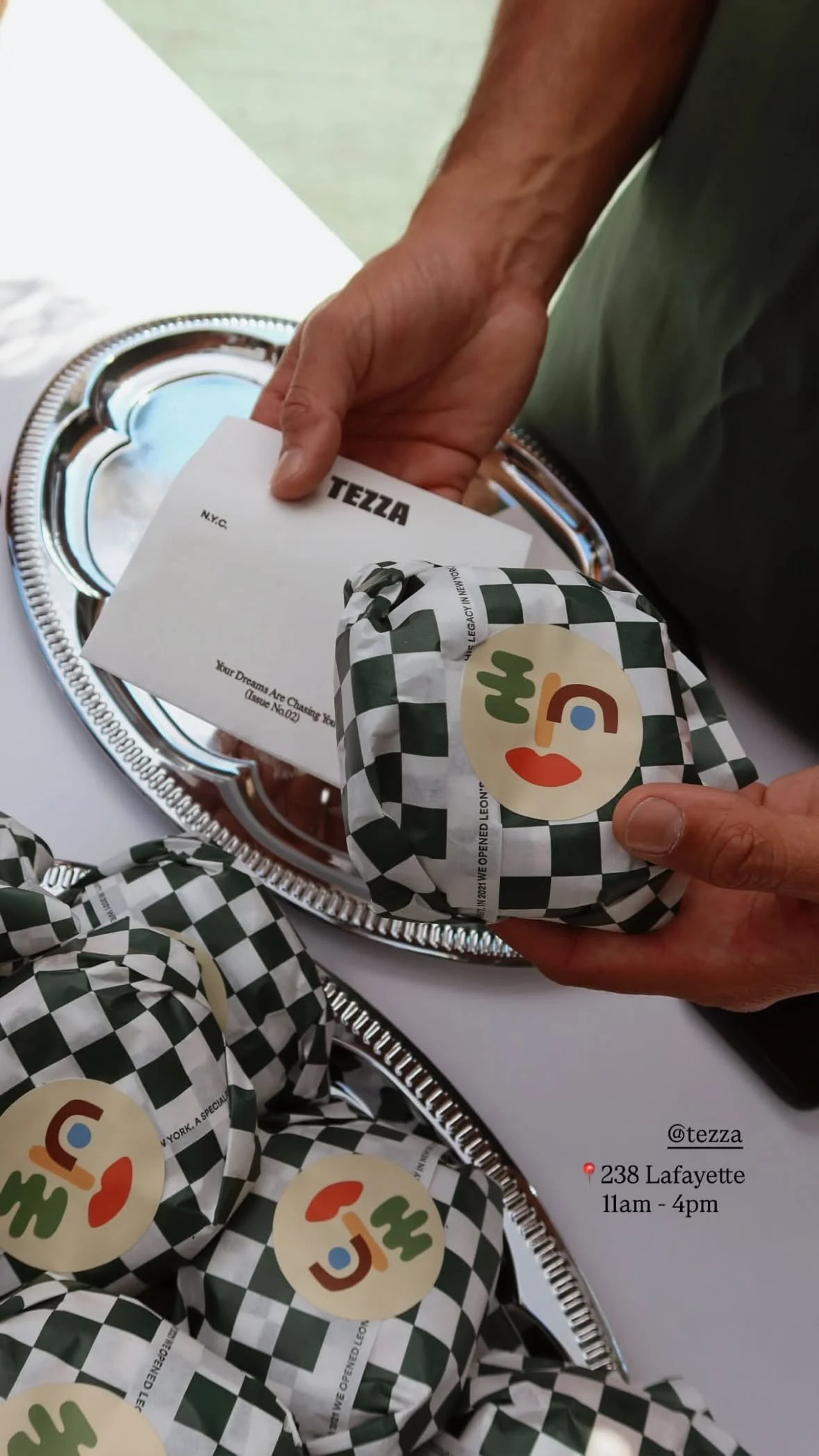 Person holding wrapped food items with a sticker face, standing next to a silver tray with more wrapped items, with a paper that says 'TEZZA' and address info on a table.