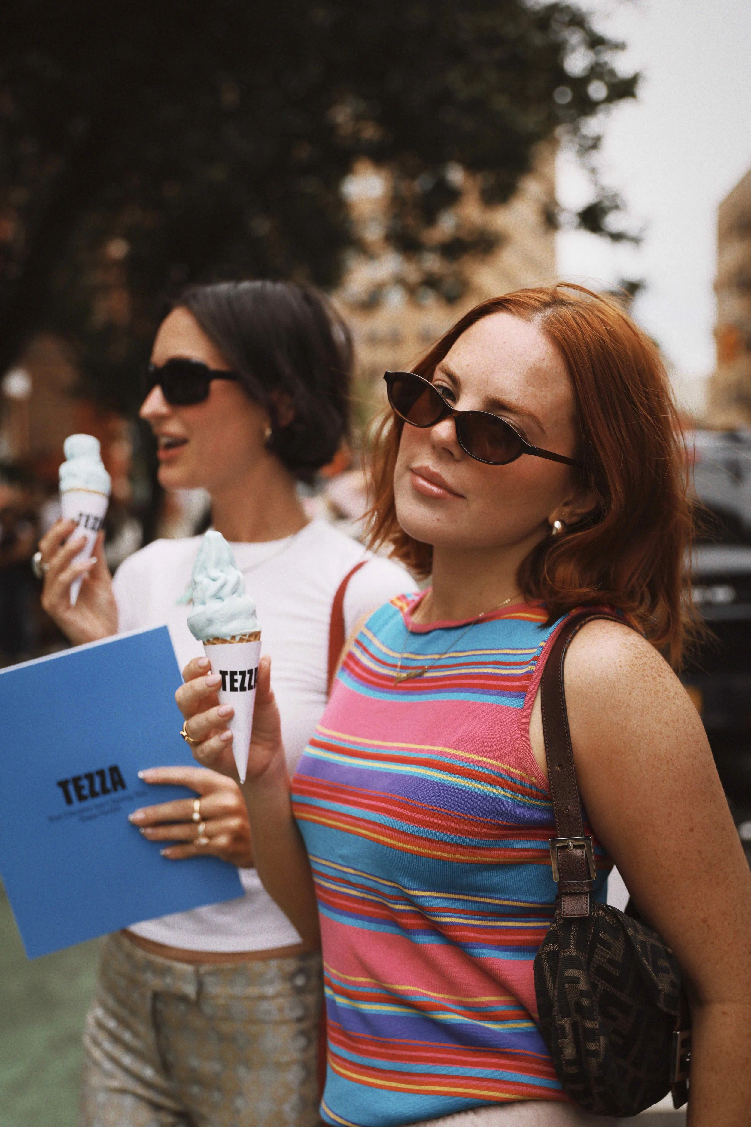 Two women with sunglasses holding ice cream cones, one woman is also holding a blue folder with the word TEZZ in it, outdoors on a city street.