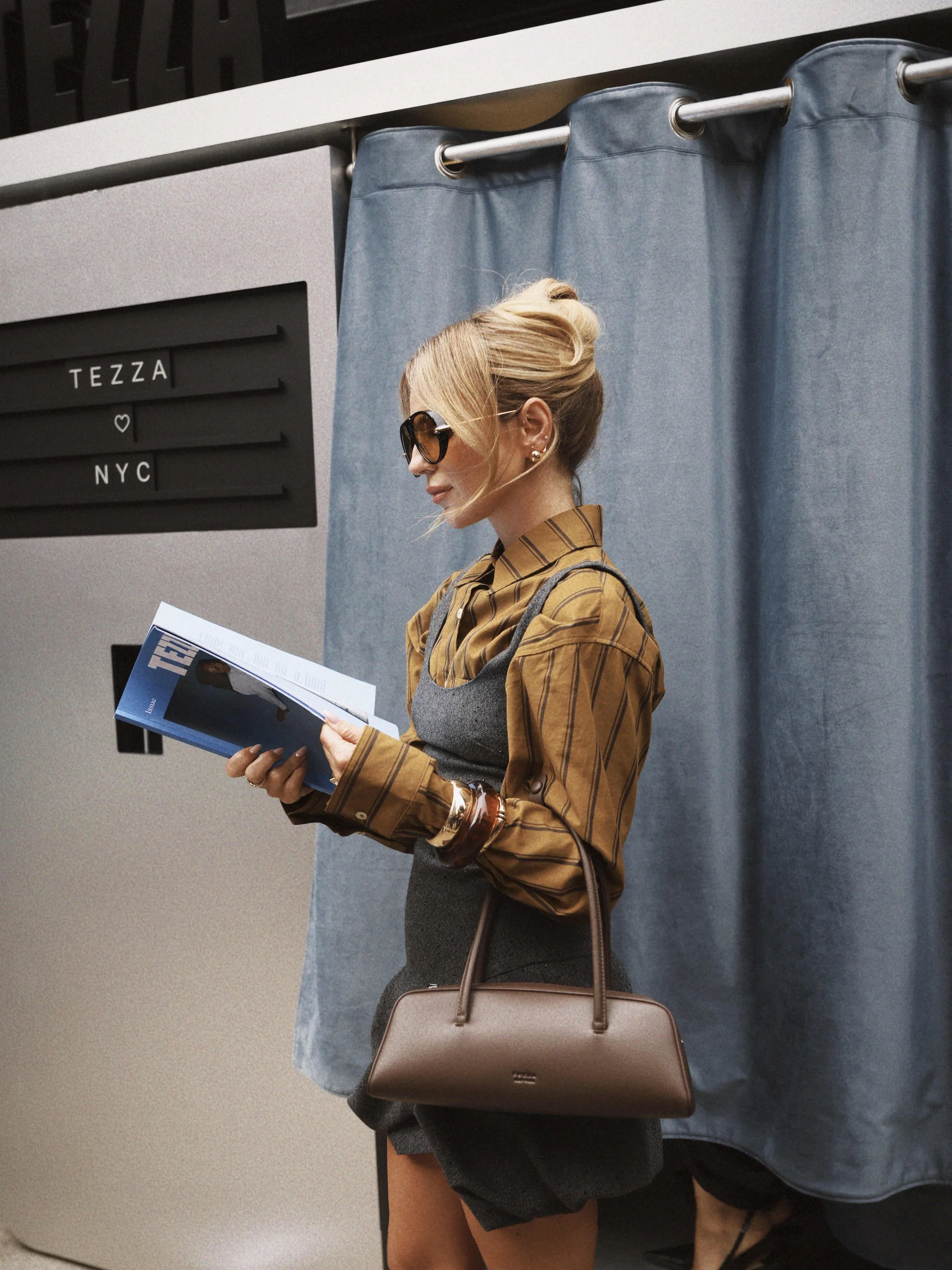 A woman with blonde hair pulled up, wearing dark sunglasses, a brown and black striped shirt under a gray dress, holding a magazine, carrying a brown handbag, standing in front of a blue curtain.