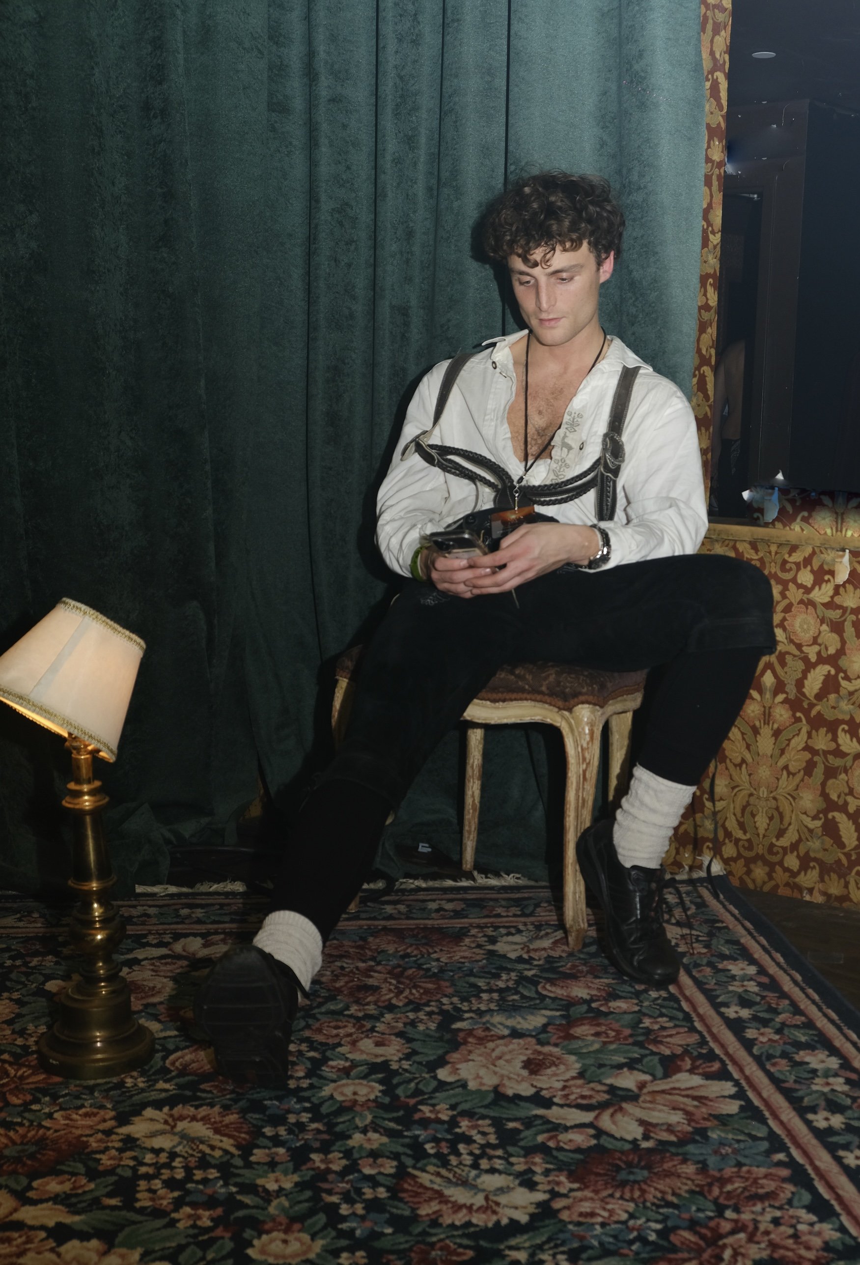 A young man with curly hair, wearing a white shirt, black pants, and black shoes, is sitting on a wooden chair, looking at his phone. The setting is indoors with dark green curtains, a patterned rug, and a small table with a lamp nearby.