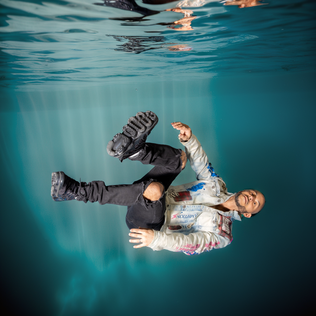 Man with beard wearing a colorful jacket and black ripped jeans floating underwater, smiling and looking at the camera.