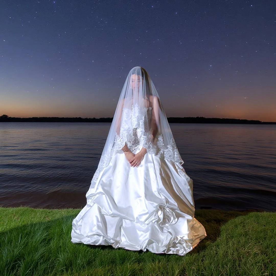 A woman in a white wedding dress with lace details, standing on grass by a body of water at dusk, with a starry sky overhead and a veil covering her face.