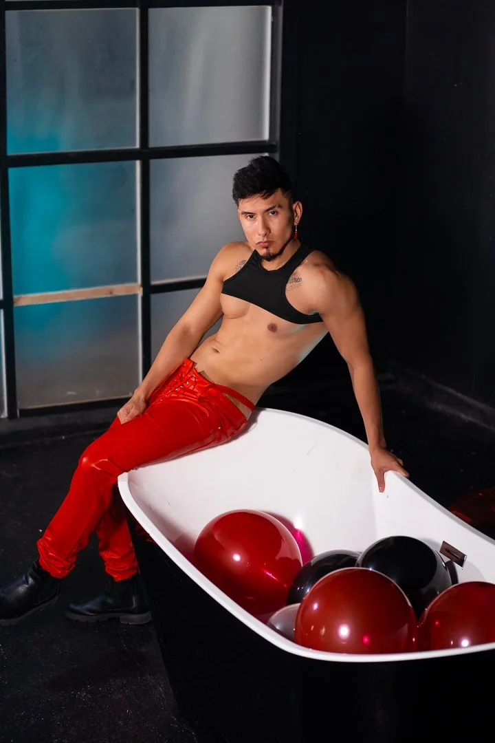 A shirtless man with dark hair and a goatee, wearing red pants and black boots, is sitting on the edge of a white bathtub filled with red and black balloons, in a dark room with a black wall and a window with frosted glass.