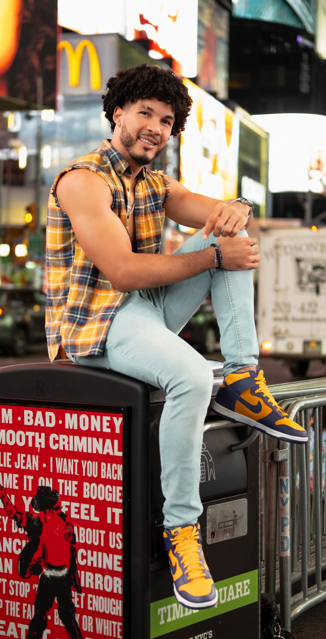 A young man with curly hair, beard, and a plaid sleeveless shirt, sitting on a NYC street island, smiling at the camera, with bright lights and billboards in Times Square behind him. He is wearing light blue jeans and yellow and navy sneakers.