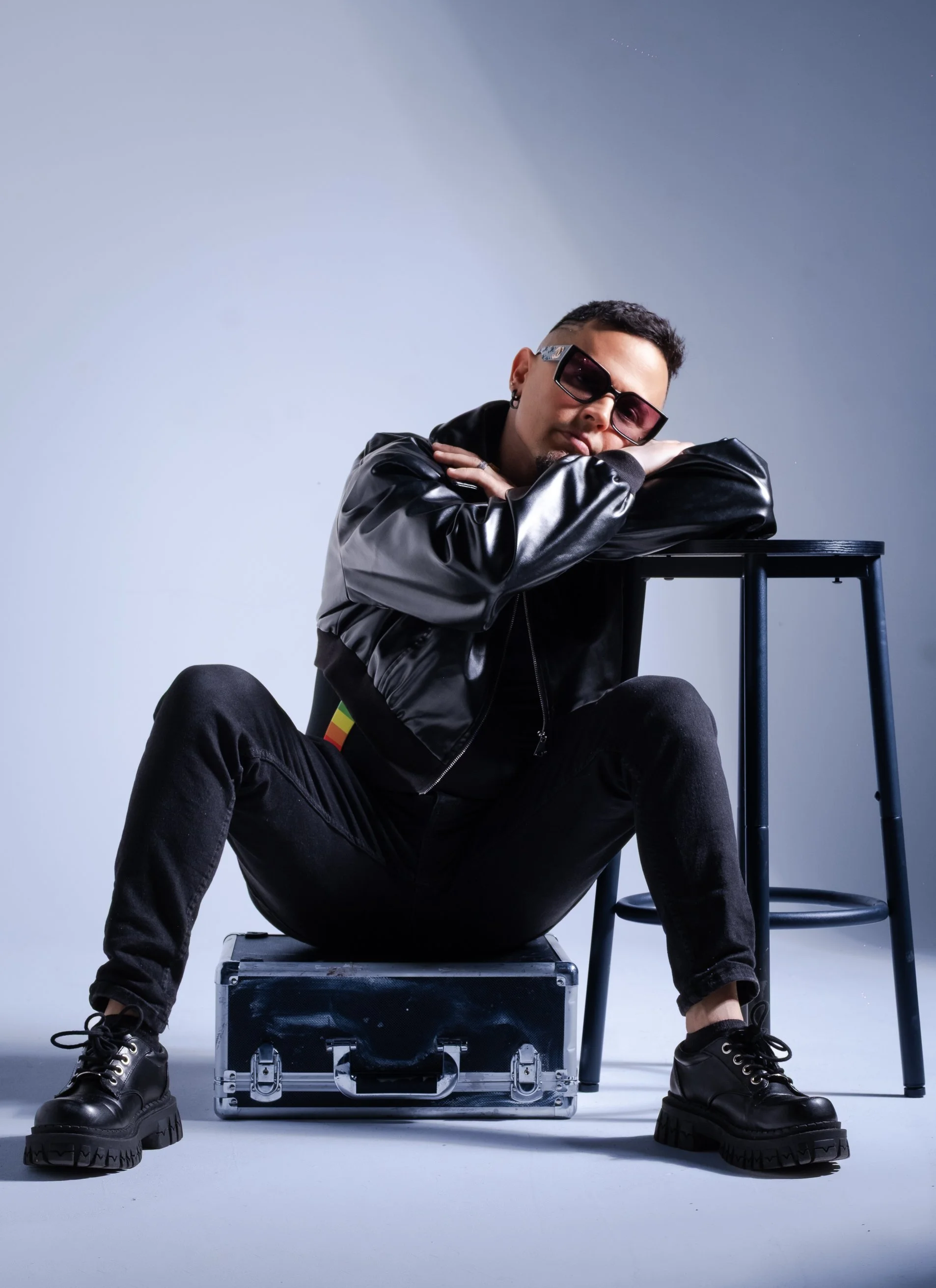 A young man with sunglasses and a black leather jacket sitting on a black box, leaning on a tall black stool with his arms crossed on the top.