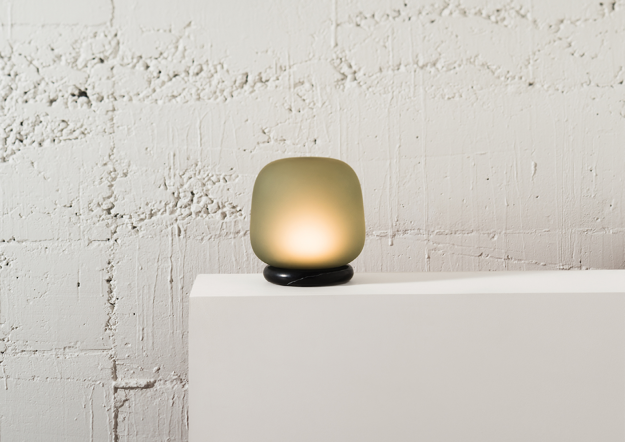 A modern table lamp with a soft, rounded, greenish frosted glass shade and a black marble base, placed on a white surface against a textured white brick wall.