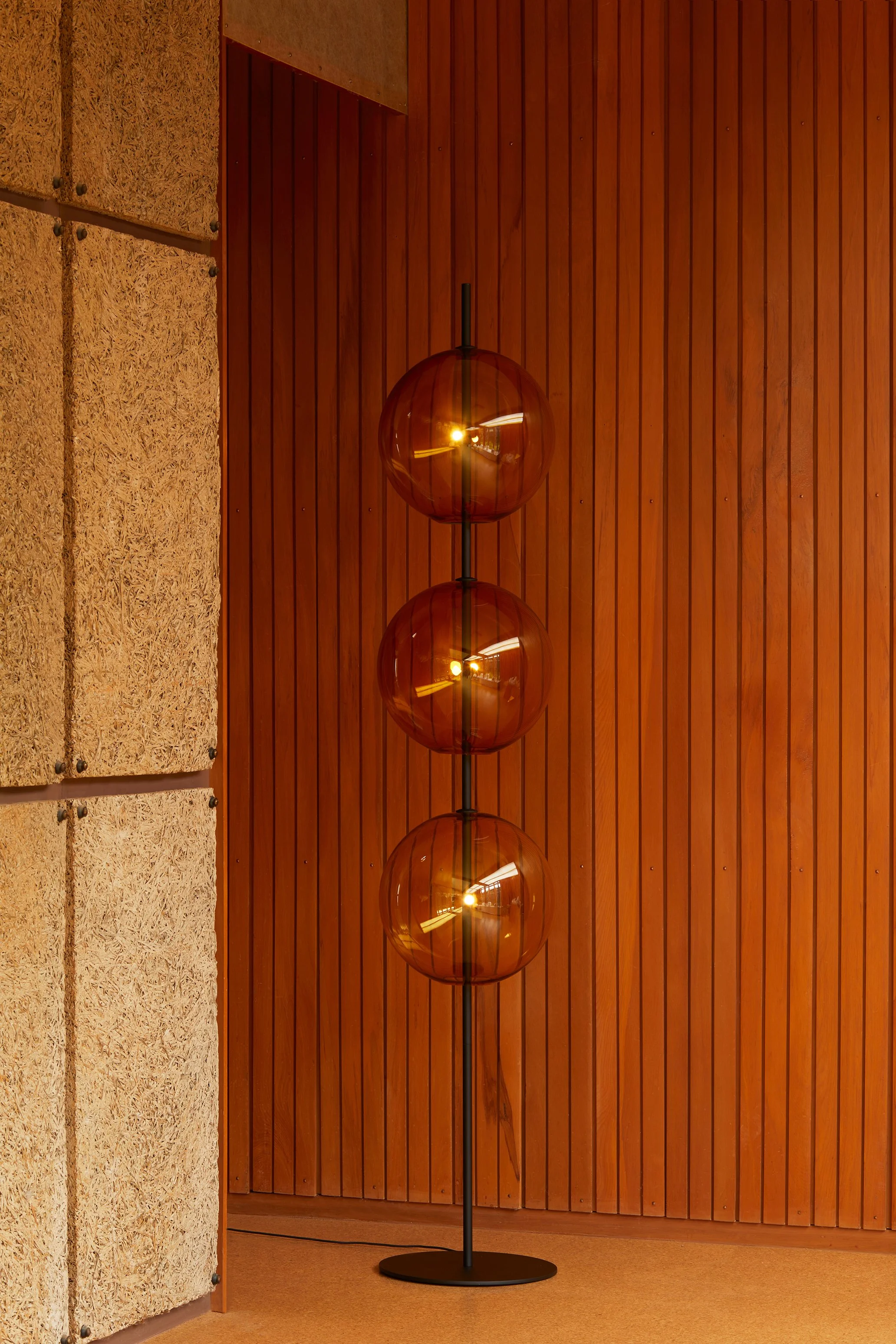 A modern floor lamp with three copper-colored glass spherical shades and warm interior lighting, standing in front of a wooden paneled wall.