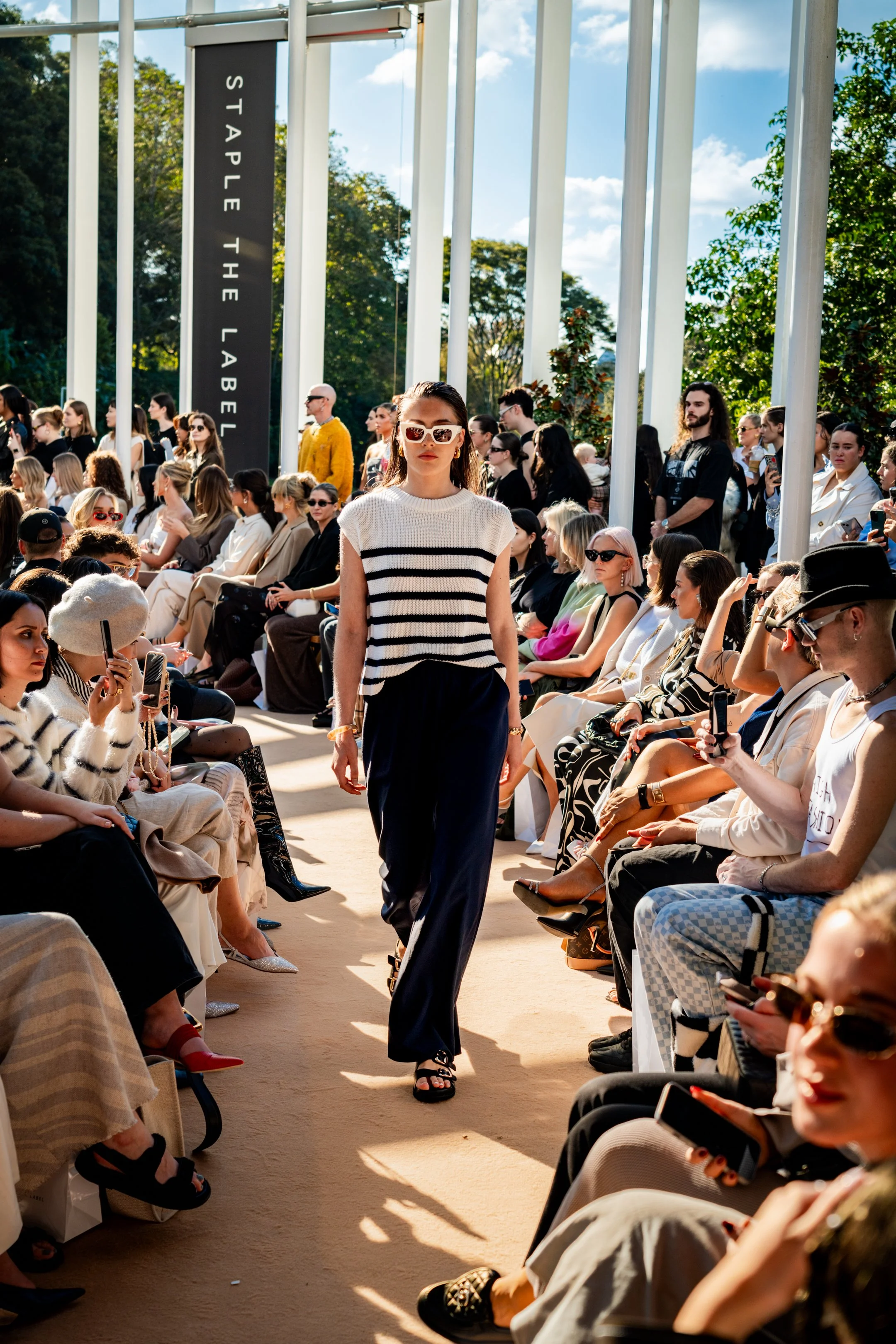 Model walking on runway at outdoor fashion show with seated audience on both sides, banners, and trees in background.