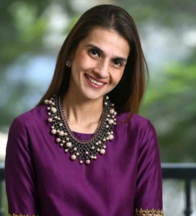 A woman wearing a purple top and a layered pearl and bead necklace, smiling with a blurred outdoor background.